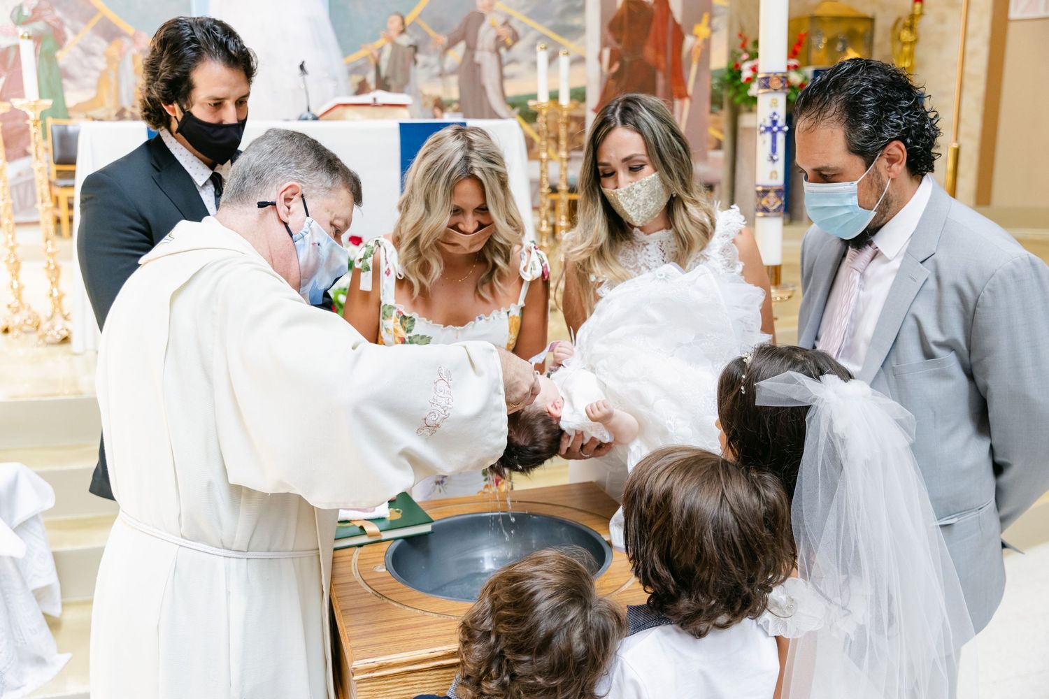 A traditional baptismal ceremony with priest and family members wearing protective masks.