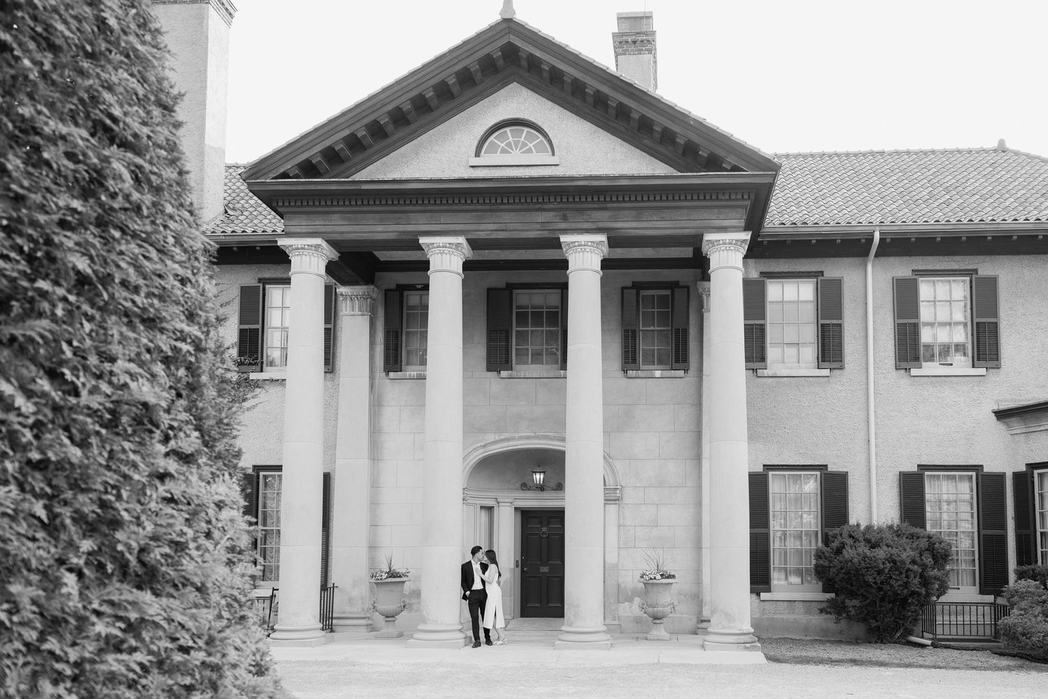 Black and white photo of historic mansion exterior with columned entrance and small figures.