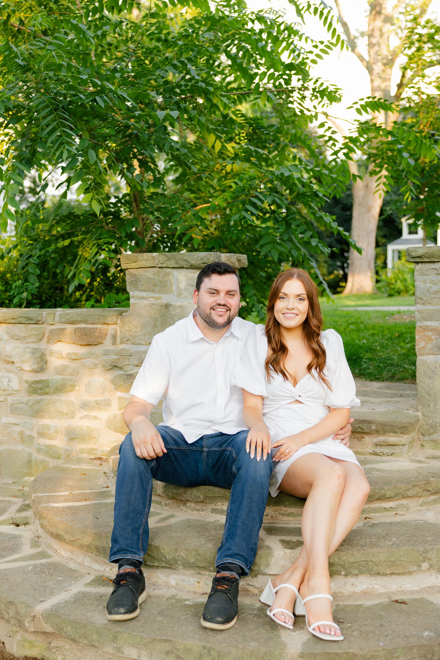 Happy couple in coordinated white outfits sitting on stone steps surrounded by summer greenery.