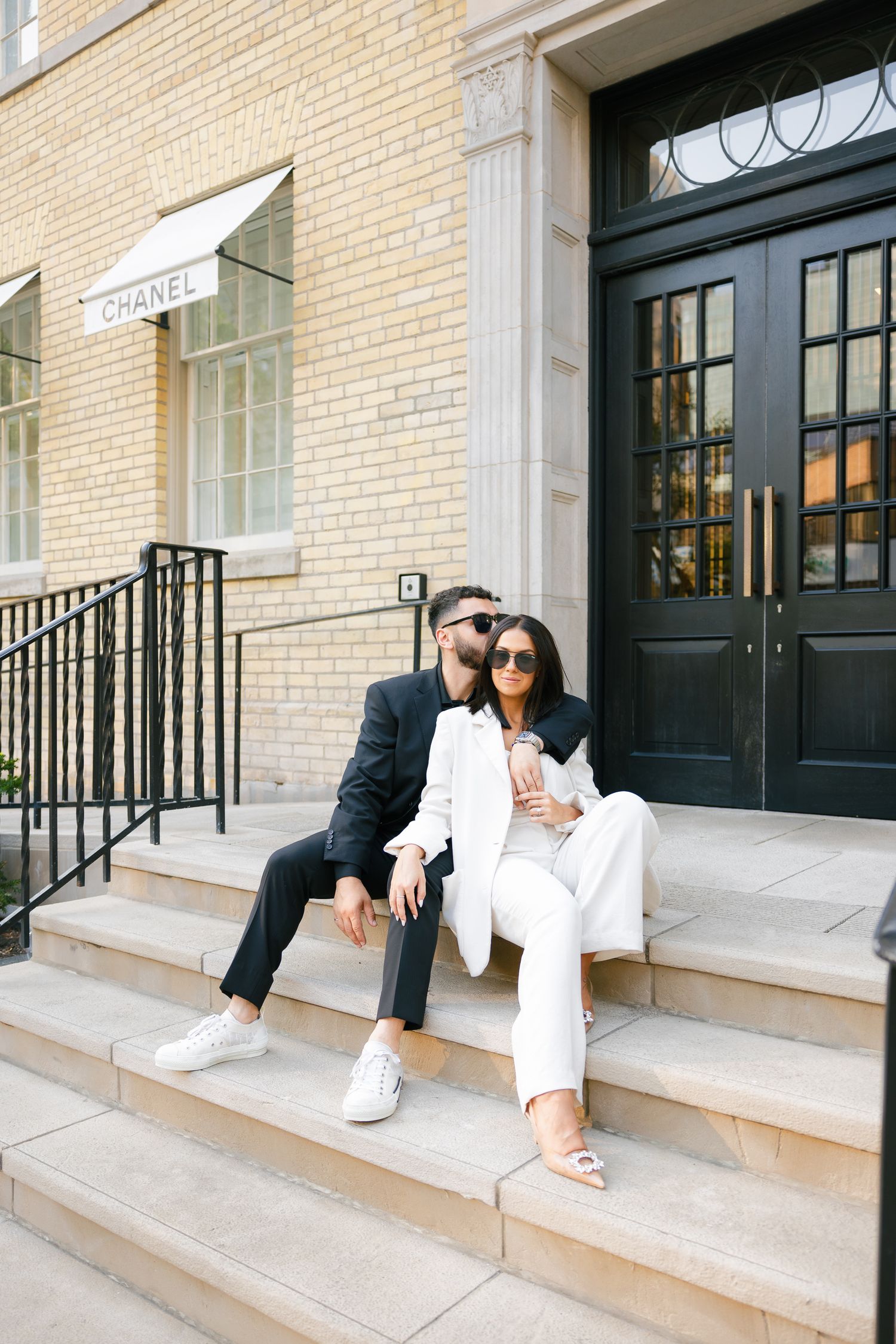 Elegant couple in black and white attire lounging on steps of Chanel boutique.