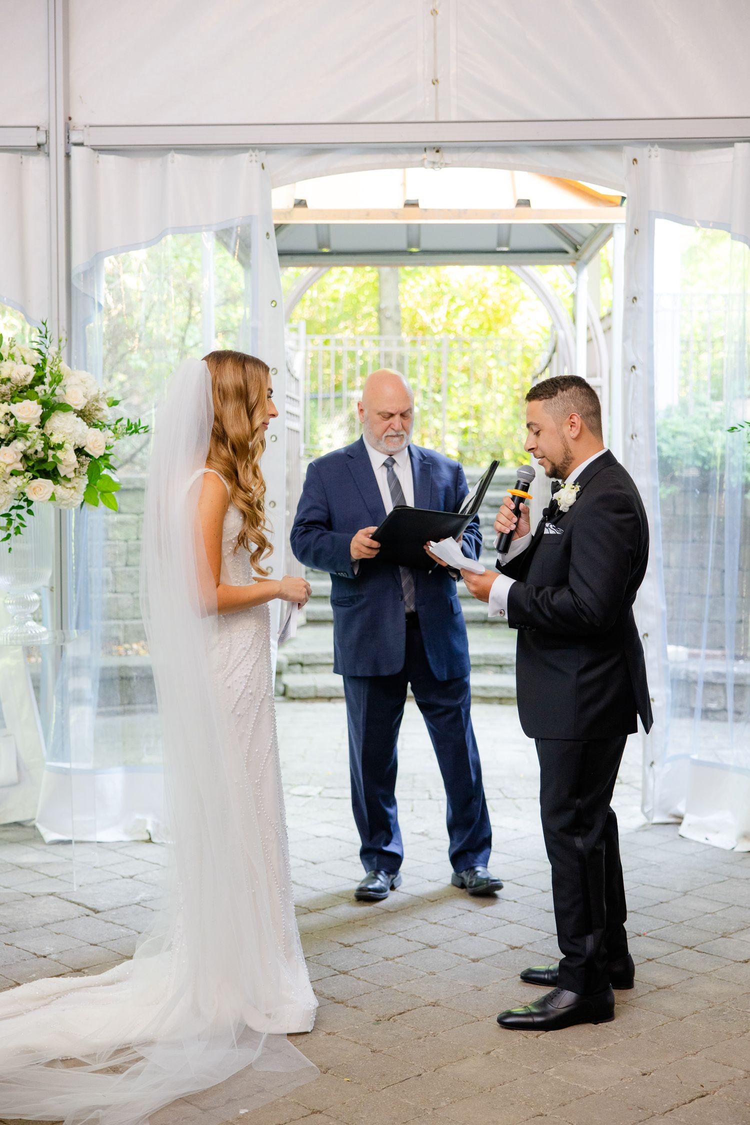 Wedding ceremony with officiant and couple exchanging vows under white tent with floral decor.