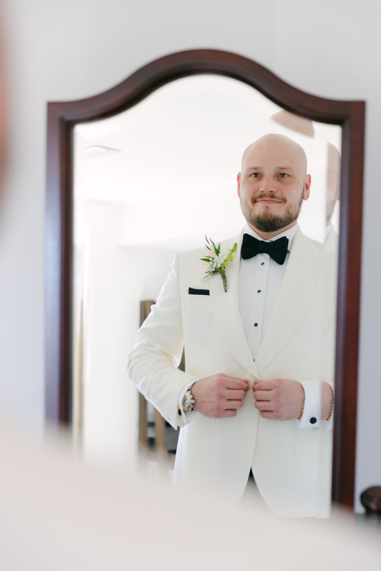 A reflection in a wooden framed mirror shows a groom in classic white tuxedo adjusting his outfit.