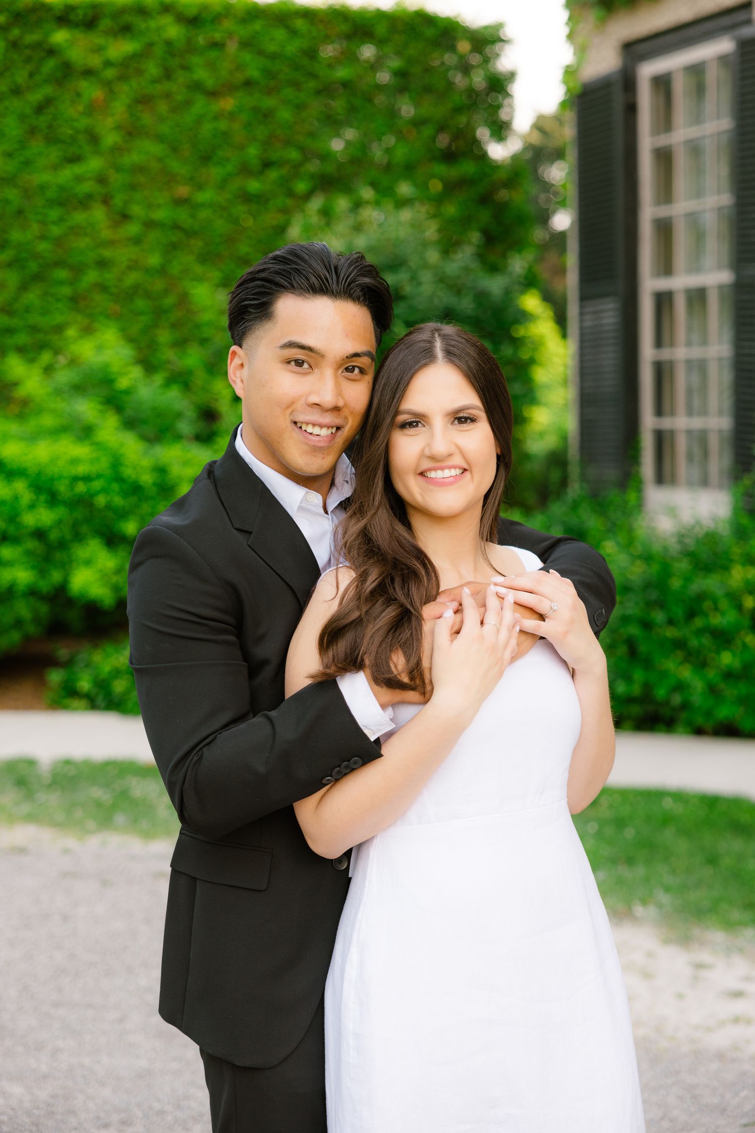 Engaged couple embraces outdoors, with bride in white dress and groom in black suit.