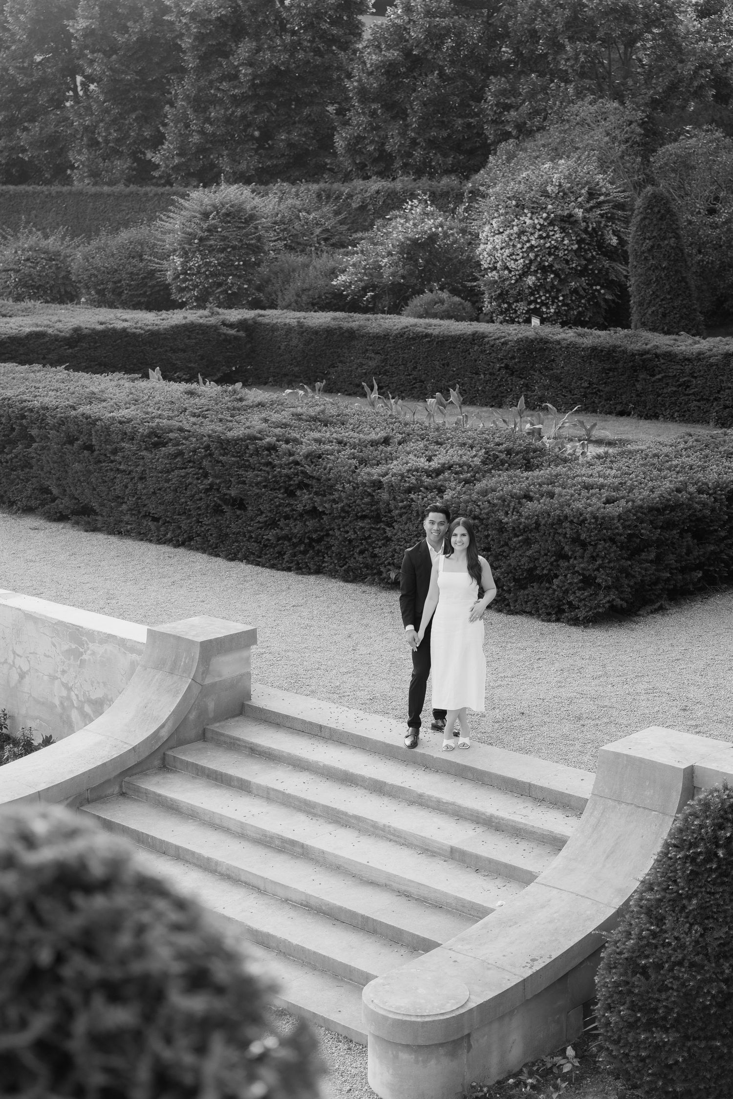 A couple in formal attire standing together on curved stone steps in a garden landscape.