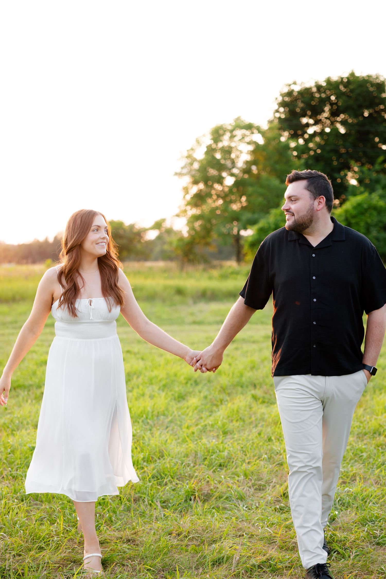 A couple holds hands while walking through a sunlit meadow, wearing contrasting black and white outfits.