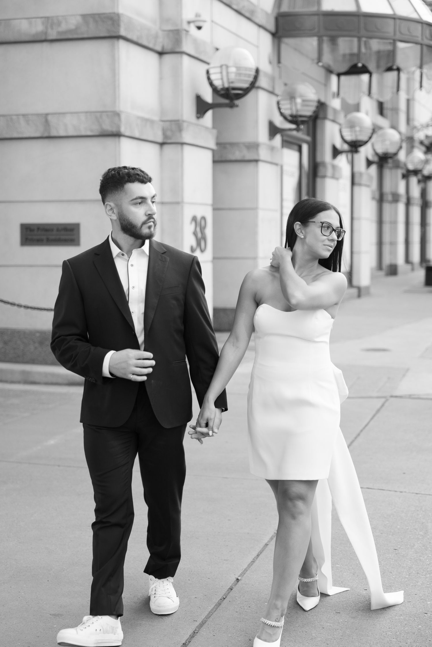 Stylish couple in formal wear walking hand in hand along a city street with architectural details.