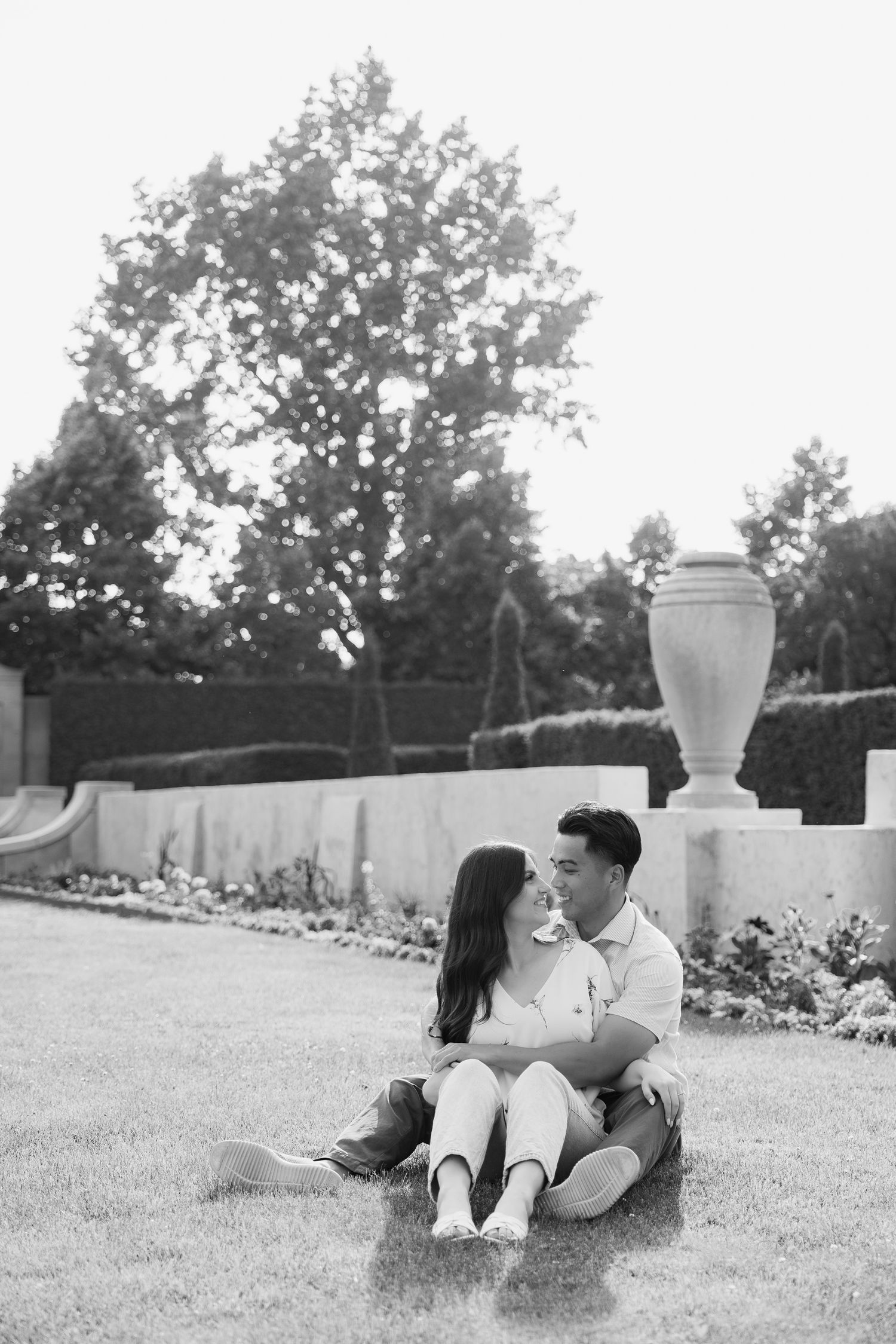 A black and white photo of a couple sharing an intimate moment on grass near a decorative urn.