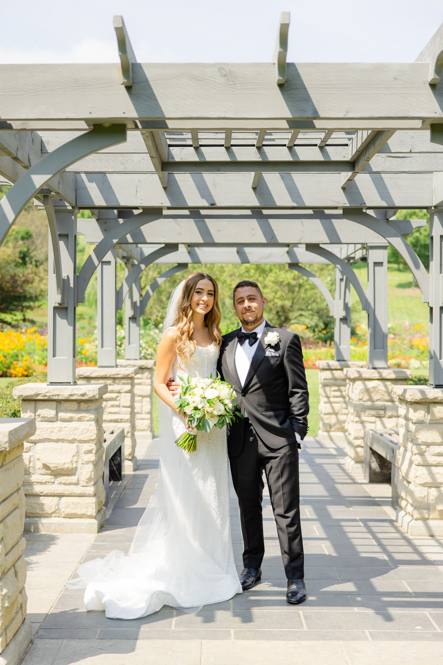 Wedding portrait of couple standing under a pergola with stone pillars and garden views.