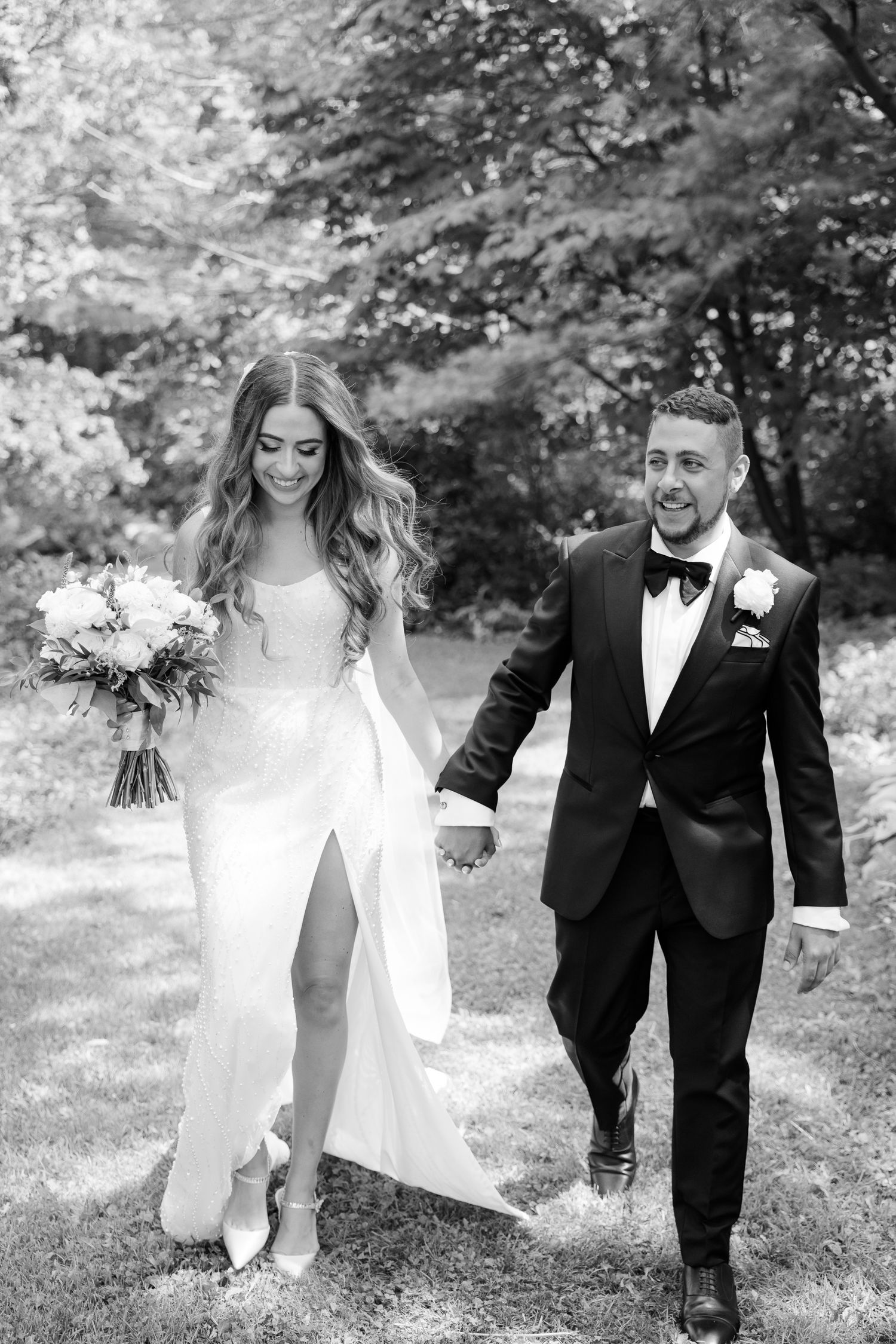 Black and white photo of newlyweds walking together holding hands in an outdoor garden setting.