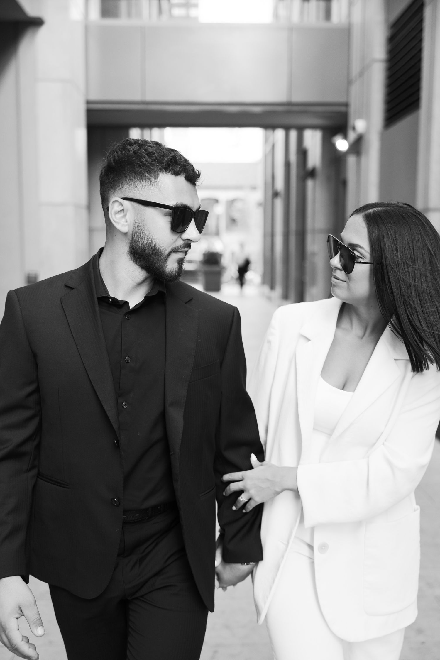 Couple in sunglasses sharing a moment while walking through a modern building corridor.