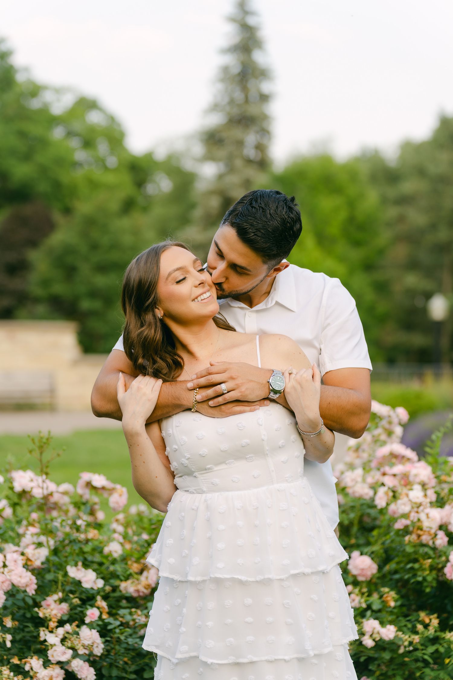 A loving couple shares an intimate moment in front of a rose garden on a sunny day.
