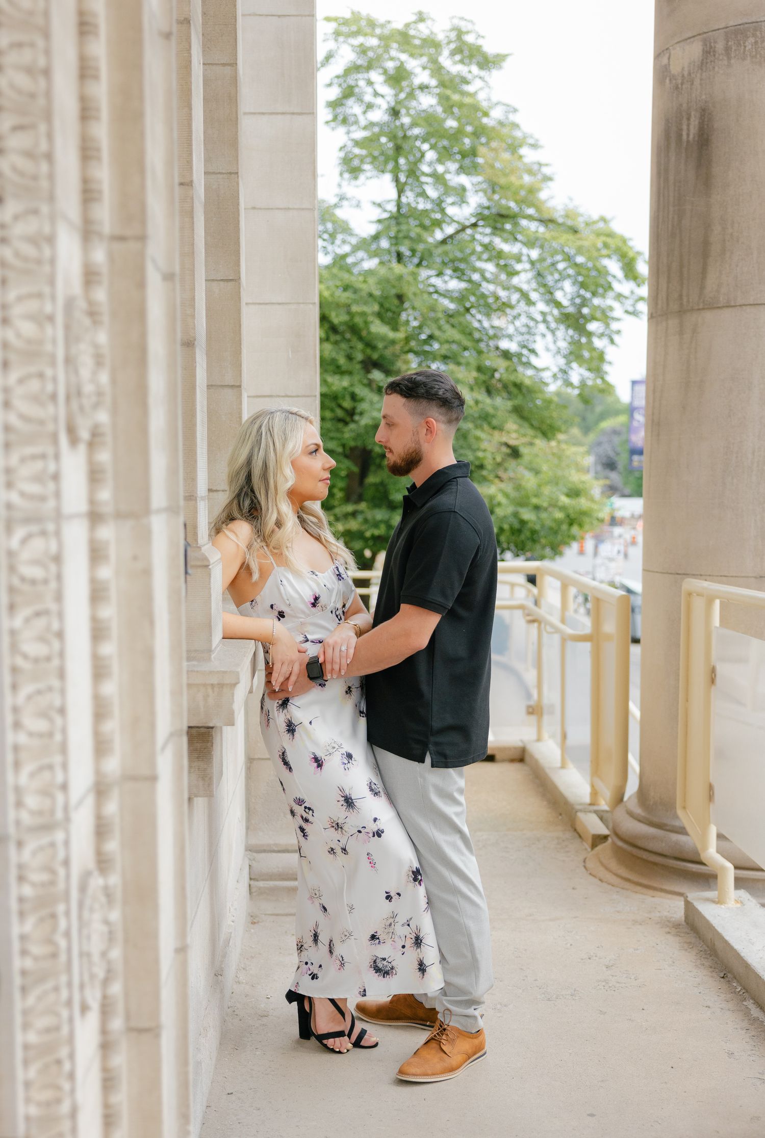 A couple stands in conversation along a columned walkway with lush greenery in background.