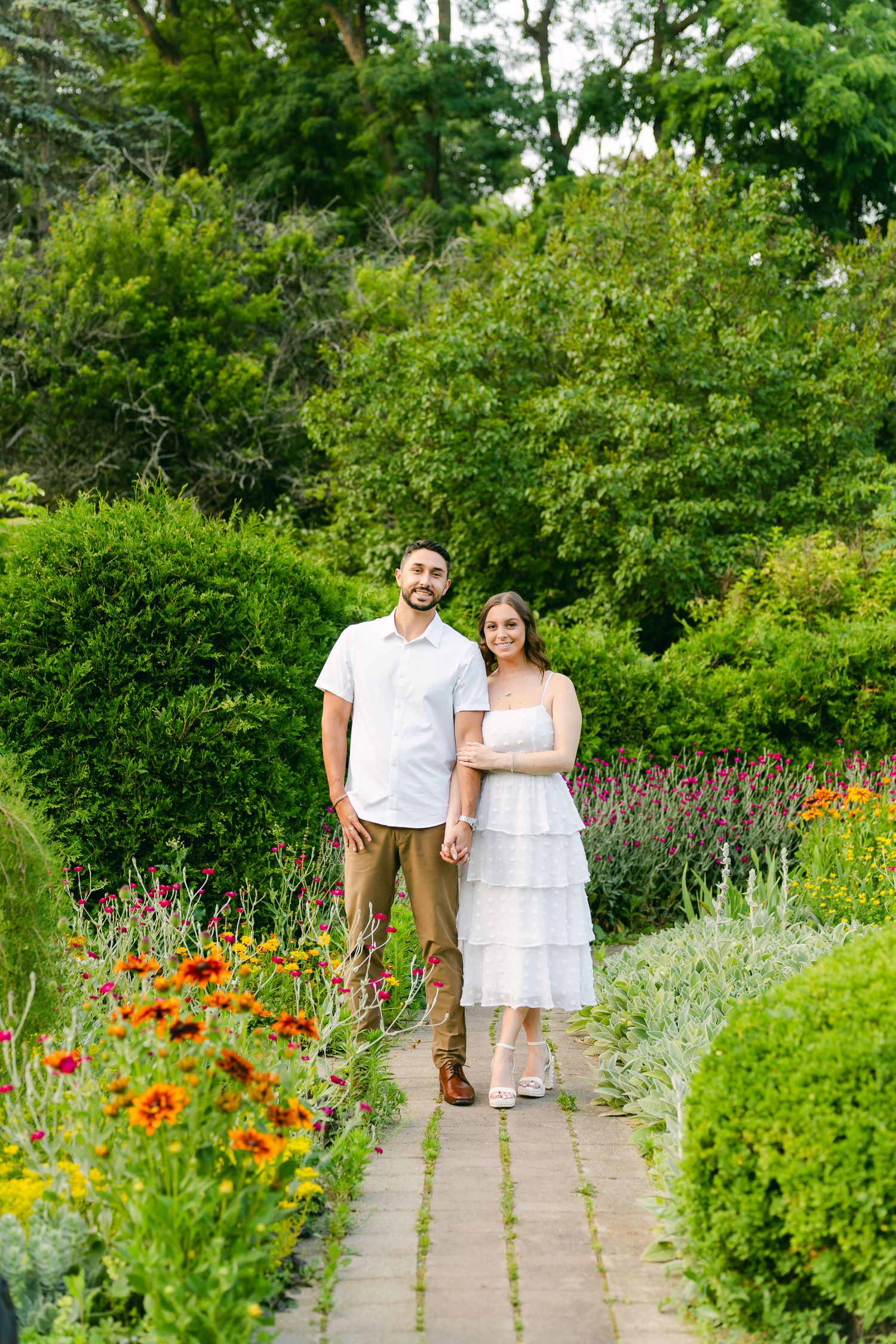 Couple walking together on garden path lined with colorful flowers and hedges.