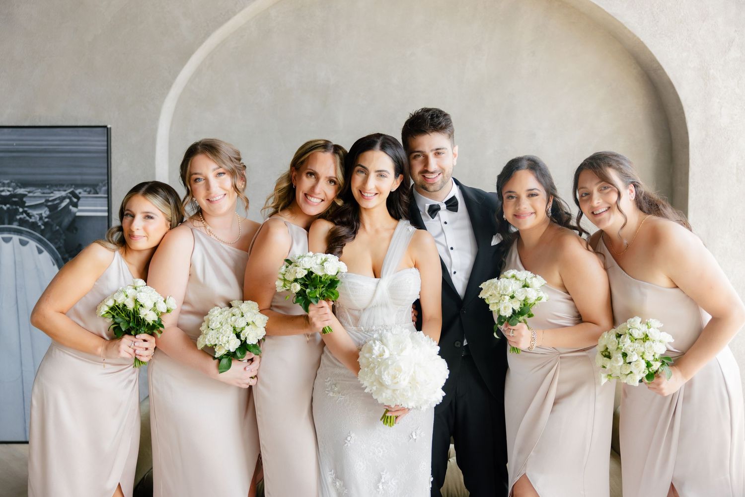 Joyful bridal party in coordinated blush dresses gathered for portrait under arch.