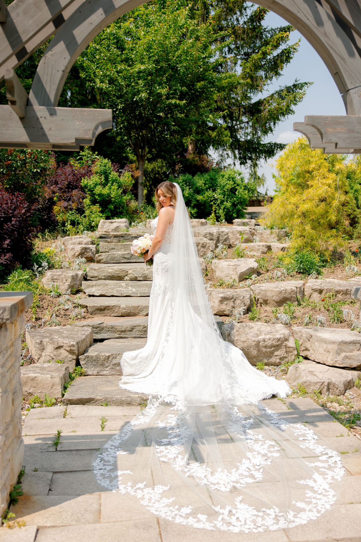 Long cathedral veil trails beautifully behind a bridal gown on landscaped stone garden steps.