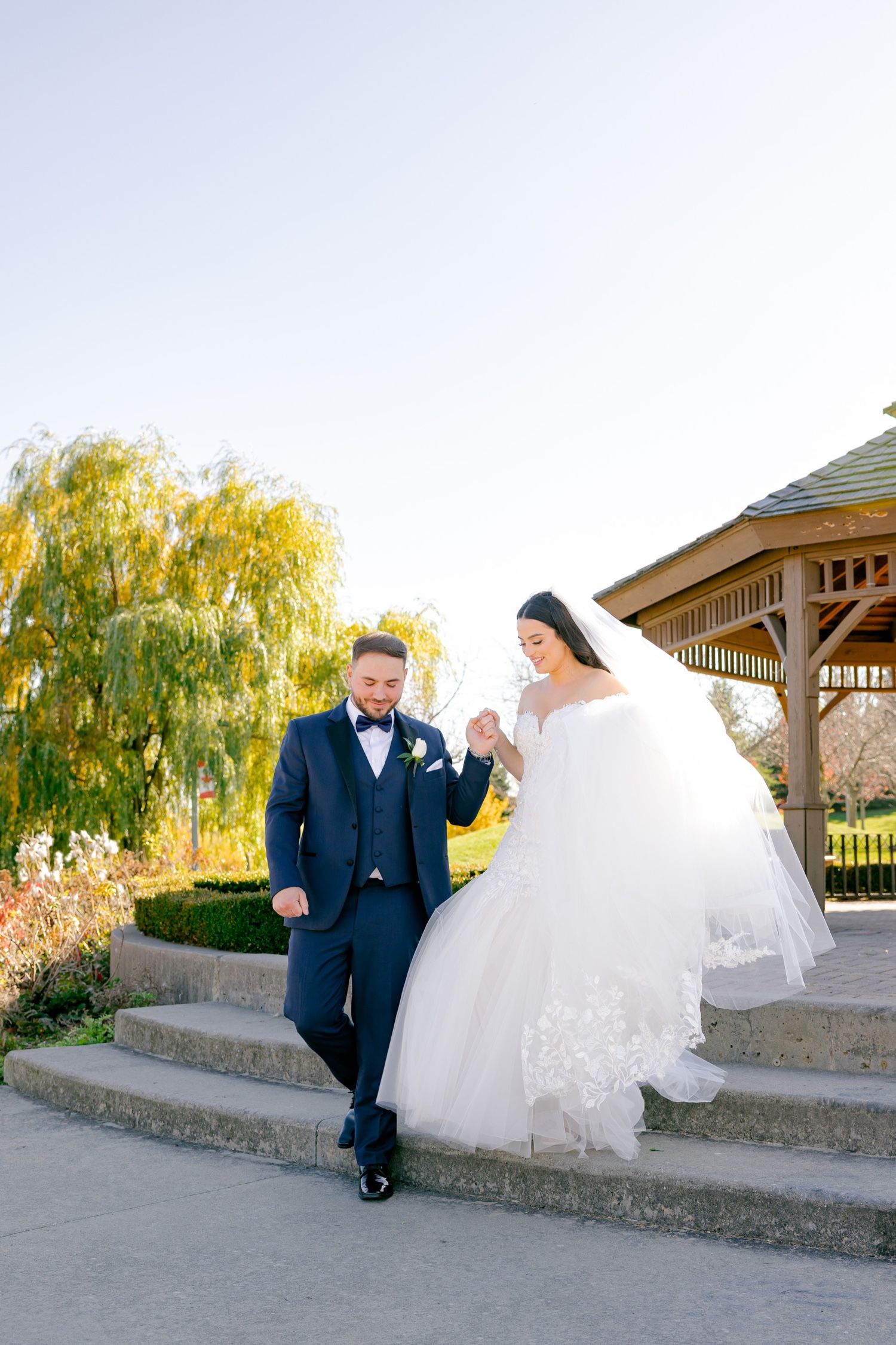 Wedding couple poses on stone steps with fall foliage and gazebo in background.
