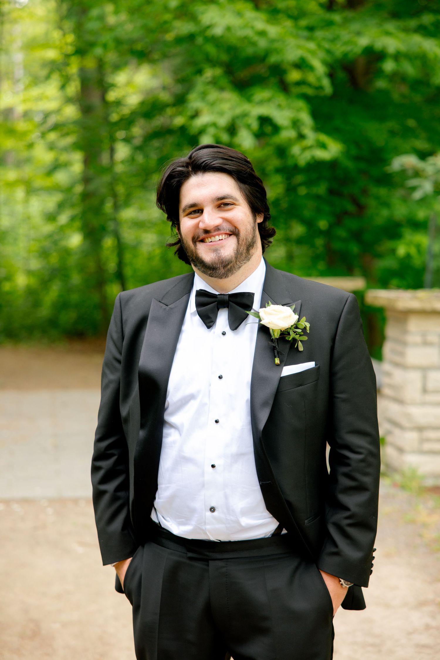 A groom in a classic black tuxedo and bowtie smiles warmly while standing outdoors against lush greenery.