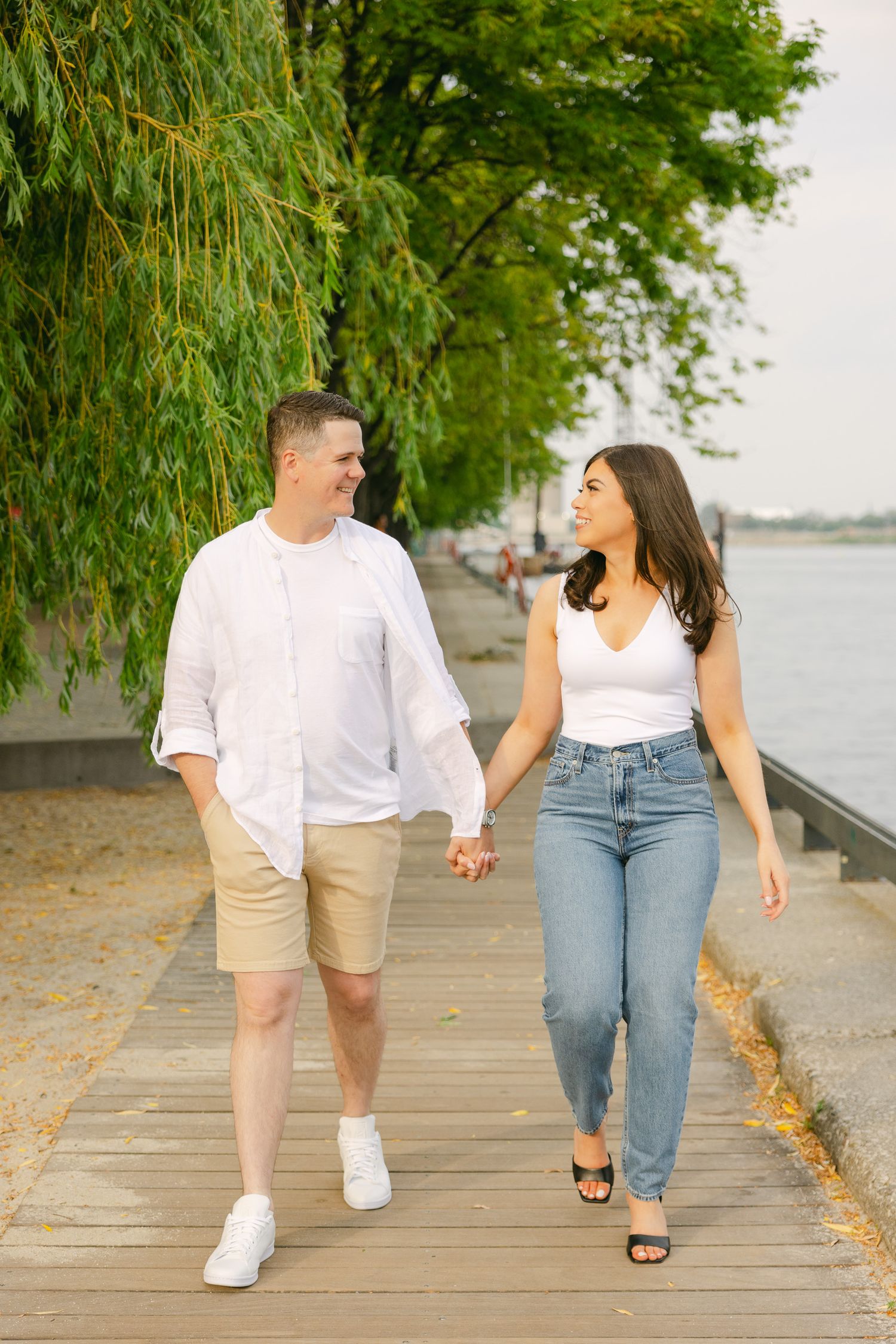 Happy couple holding hands while walking on waterfront boardwalk under willow trees.
