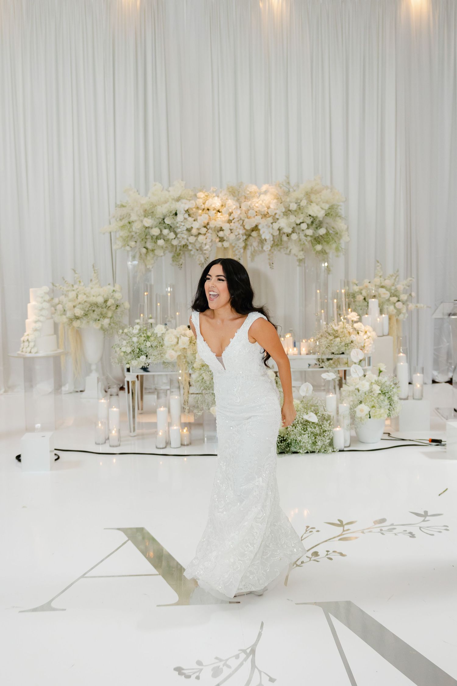 A bride poses in a fitted lace gown against an elegant white backdrop with floral arrangements.