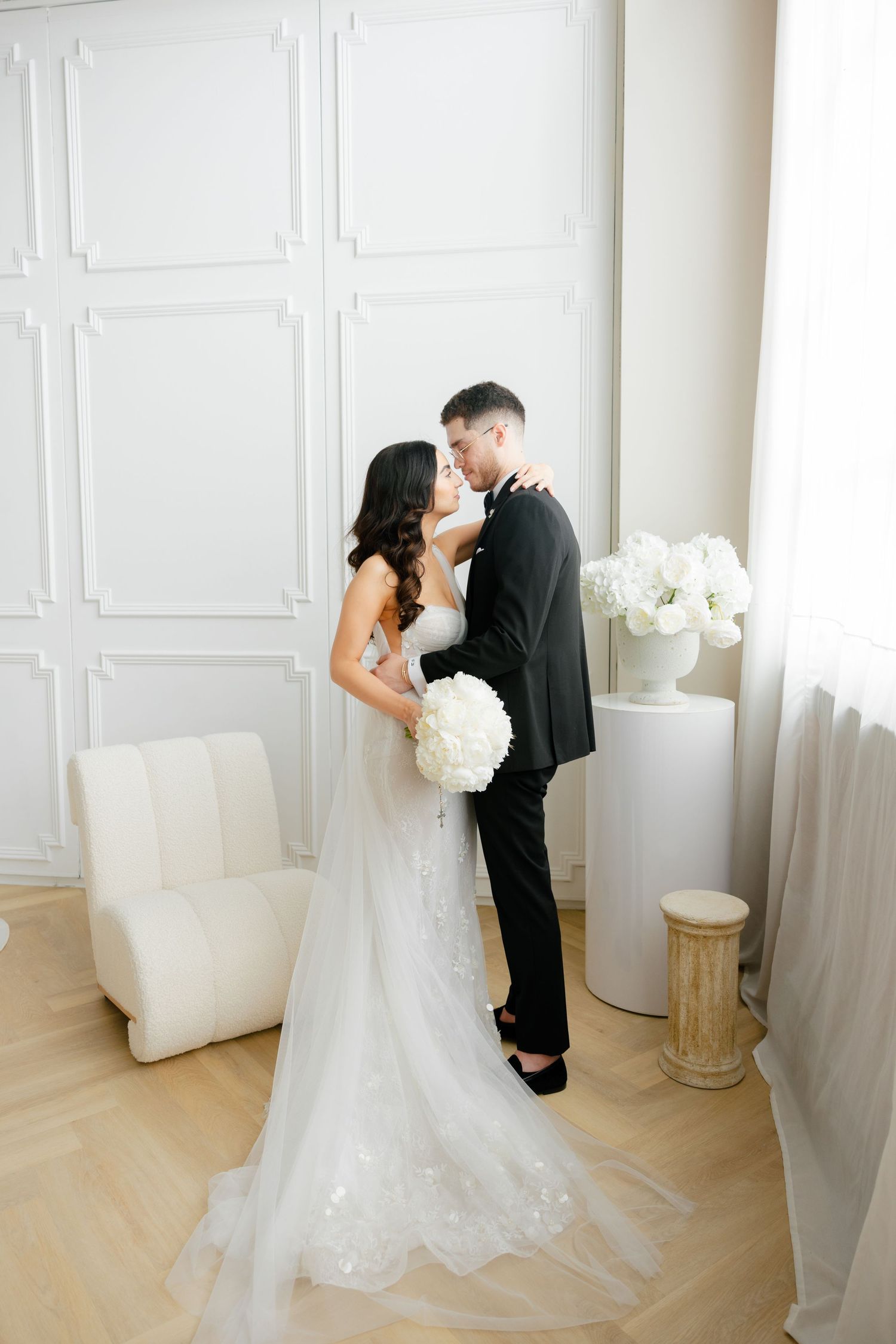 An elegant wedding couple sharing an intimate moment against white paneled walls with white floral decor.