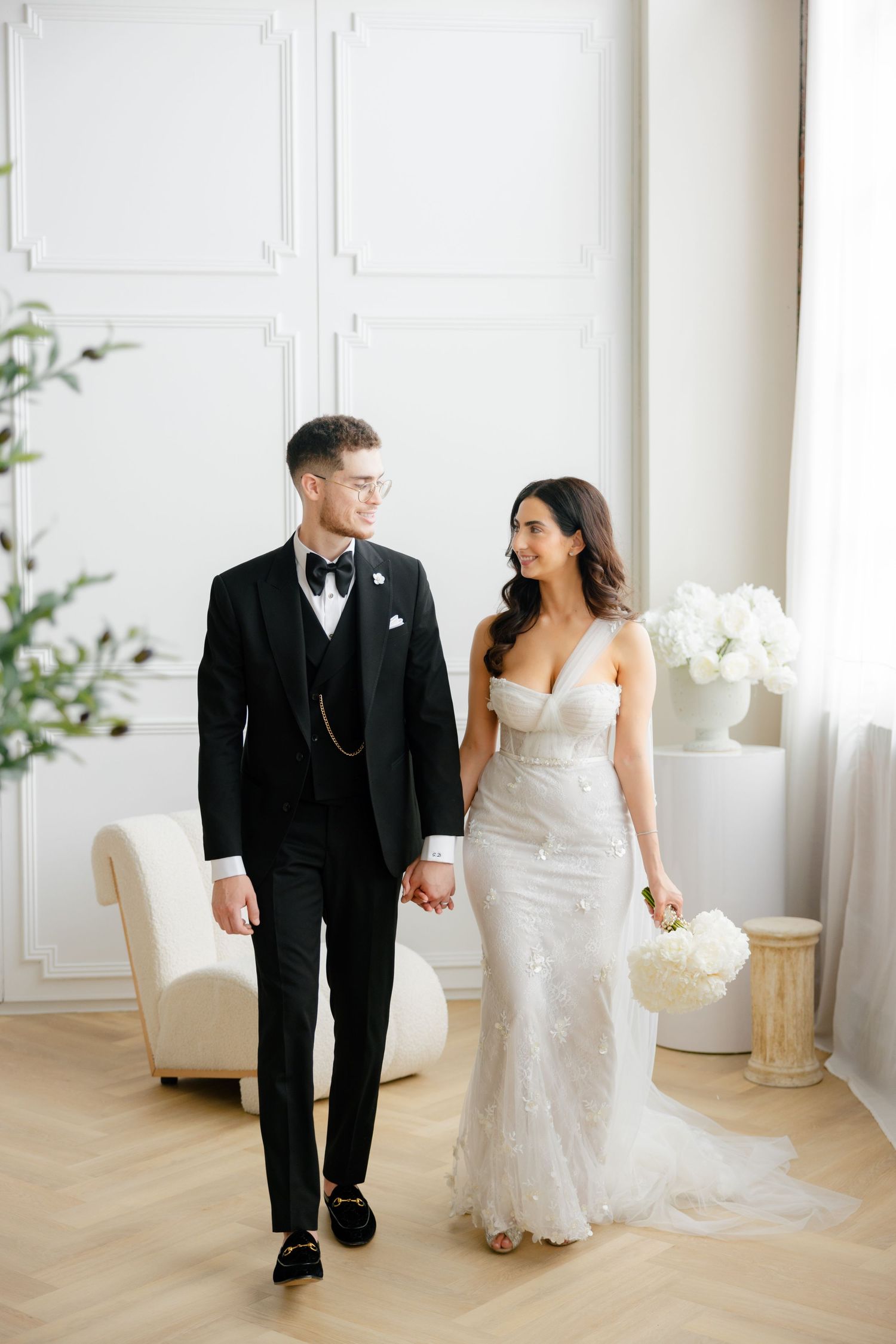 Newlyweds share a romantic moment while walking through a light-filled room with modern white furniture.