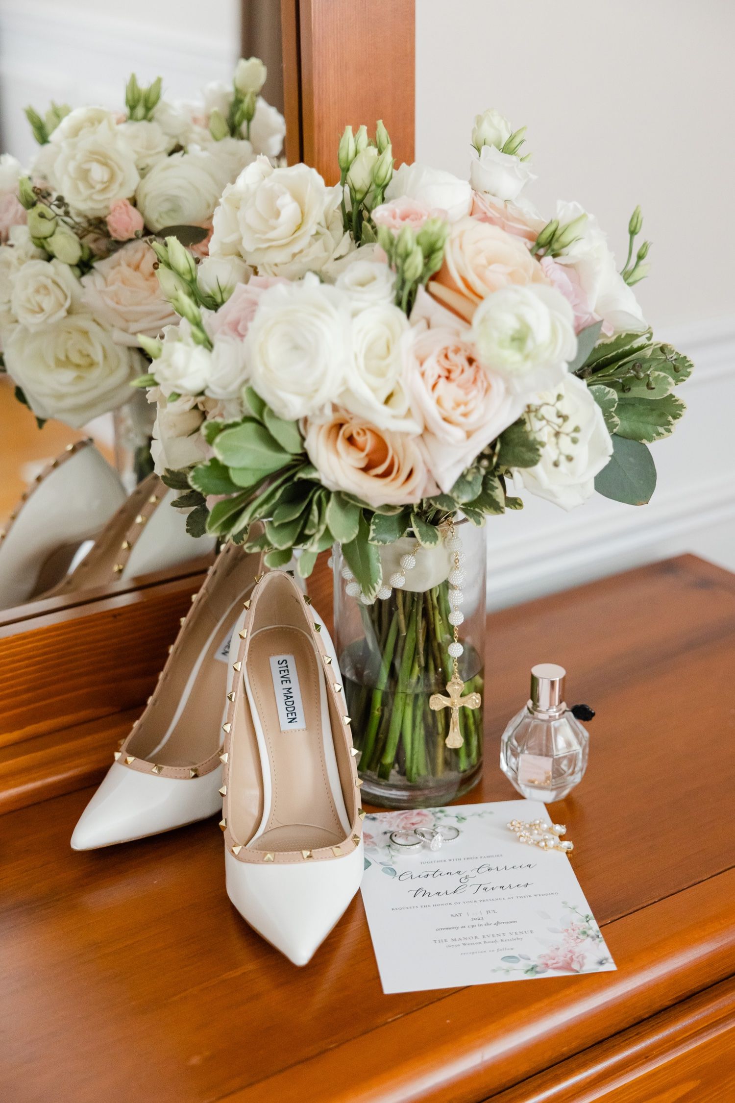 Elegant white and blush wedding bouquet with ivory roses displayed next to white heels, perfume bottle and invitation.