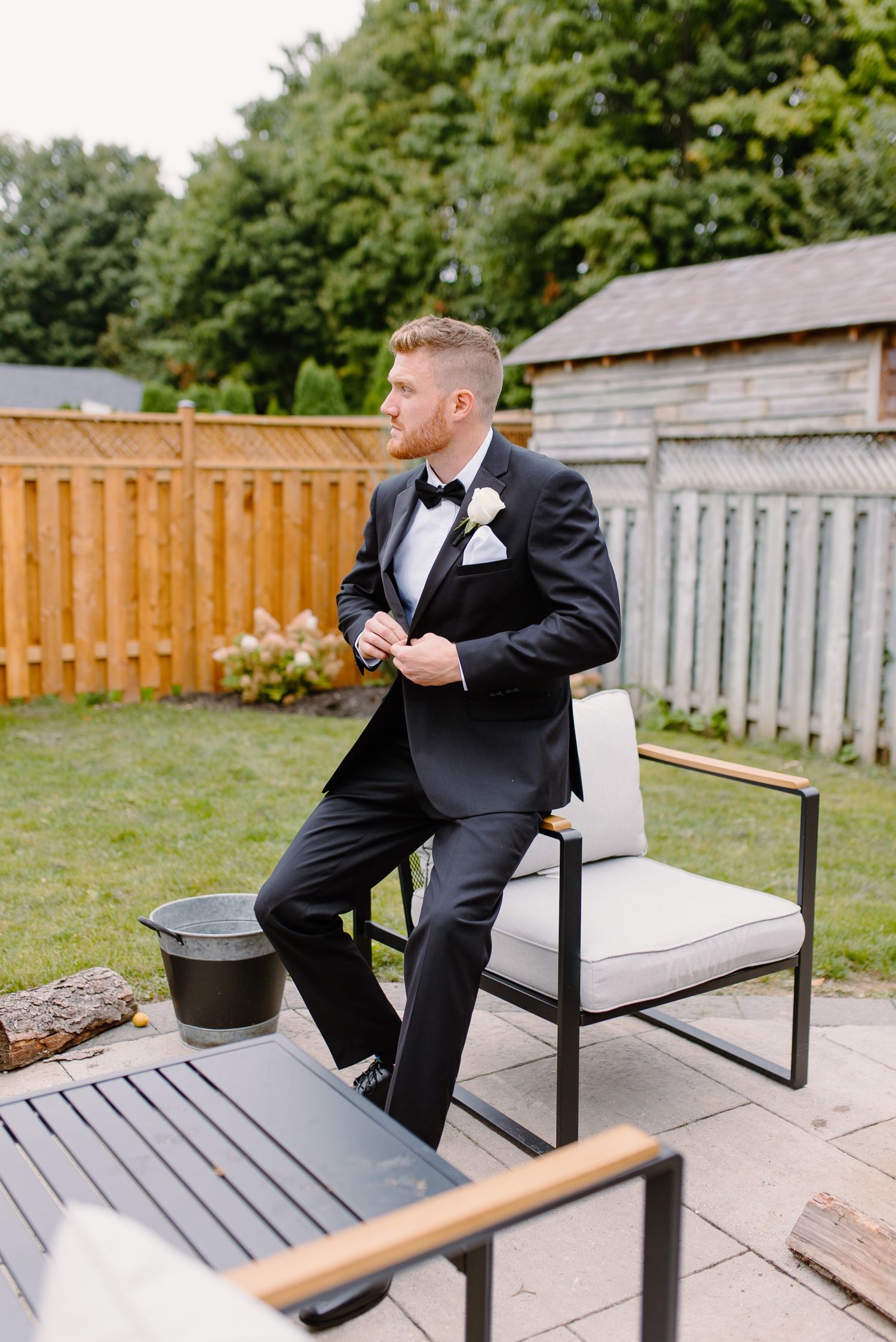 Groomsman in tuxedo sits casually on modern patio chair during outdoor photo session.