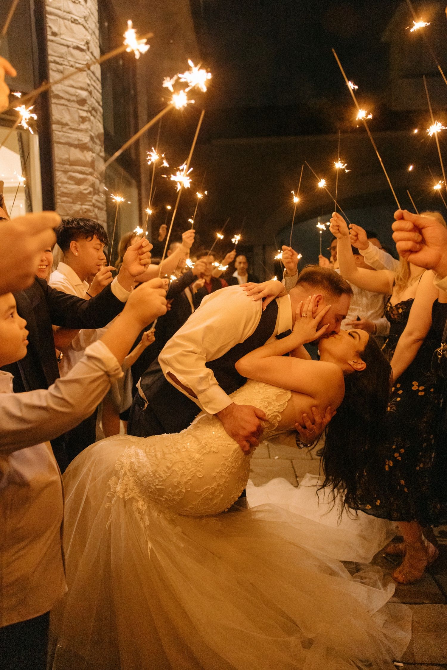 Couple shares romantic moment surrounded by guests holding sparklers at evening wedding celebration.