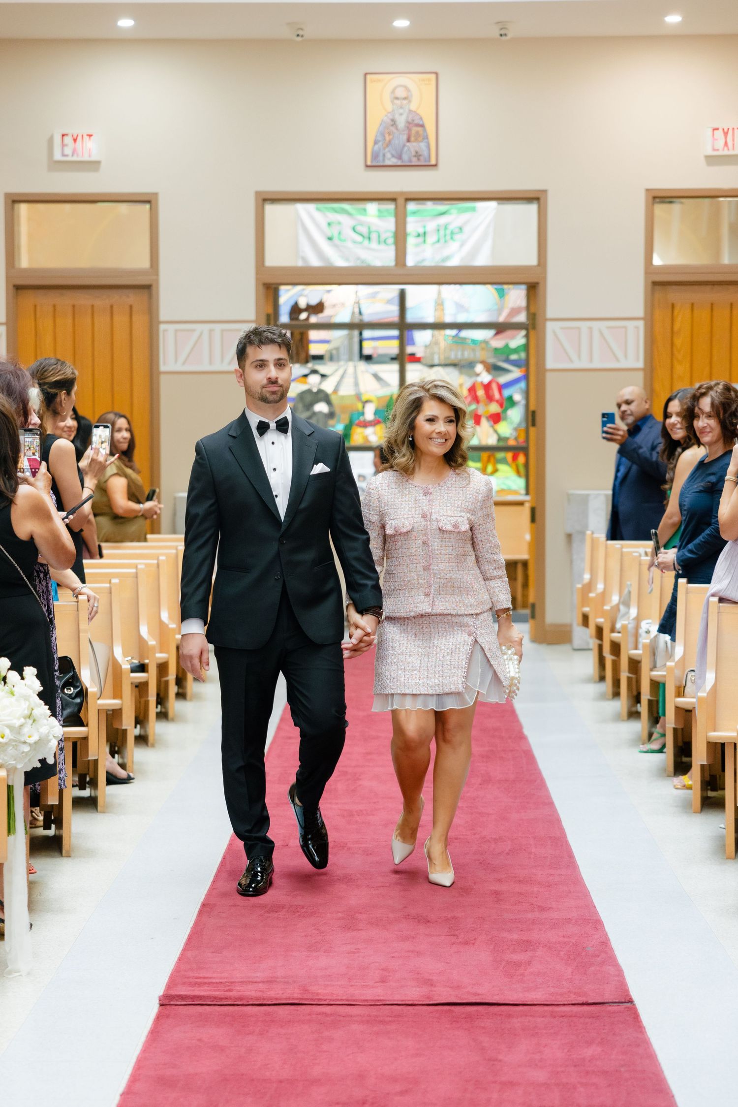Wedding party members walk down red-carpeted church aisle during traditional ceremony.