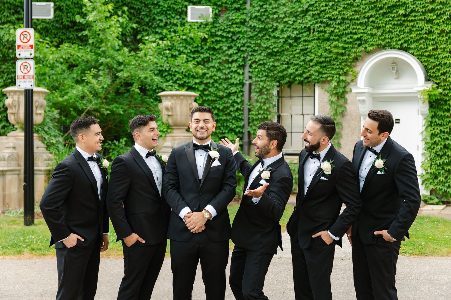 Wedding party in black tuxedos sharing a lighthearted moment against ivy-covered backdrop.