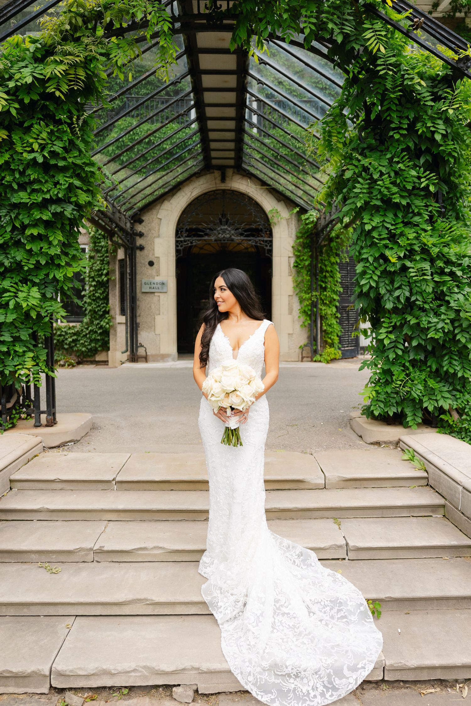 Full-length bridal portrait showing lace gown with train against greenhouse entrance covered in vines.