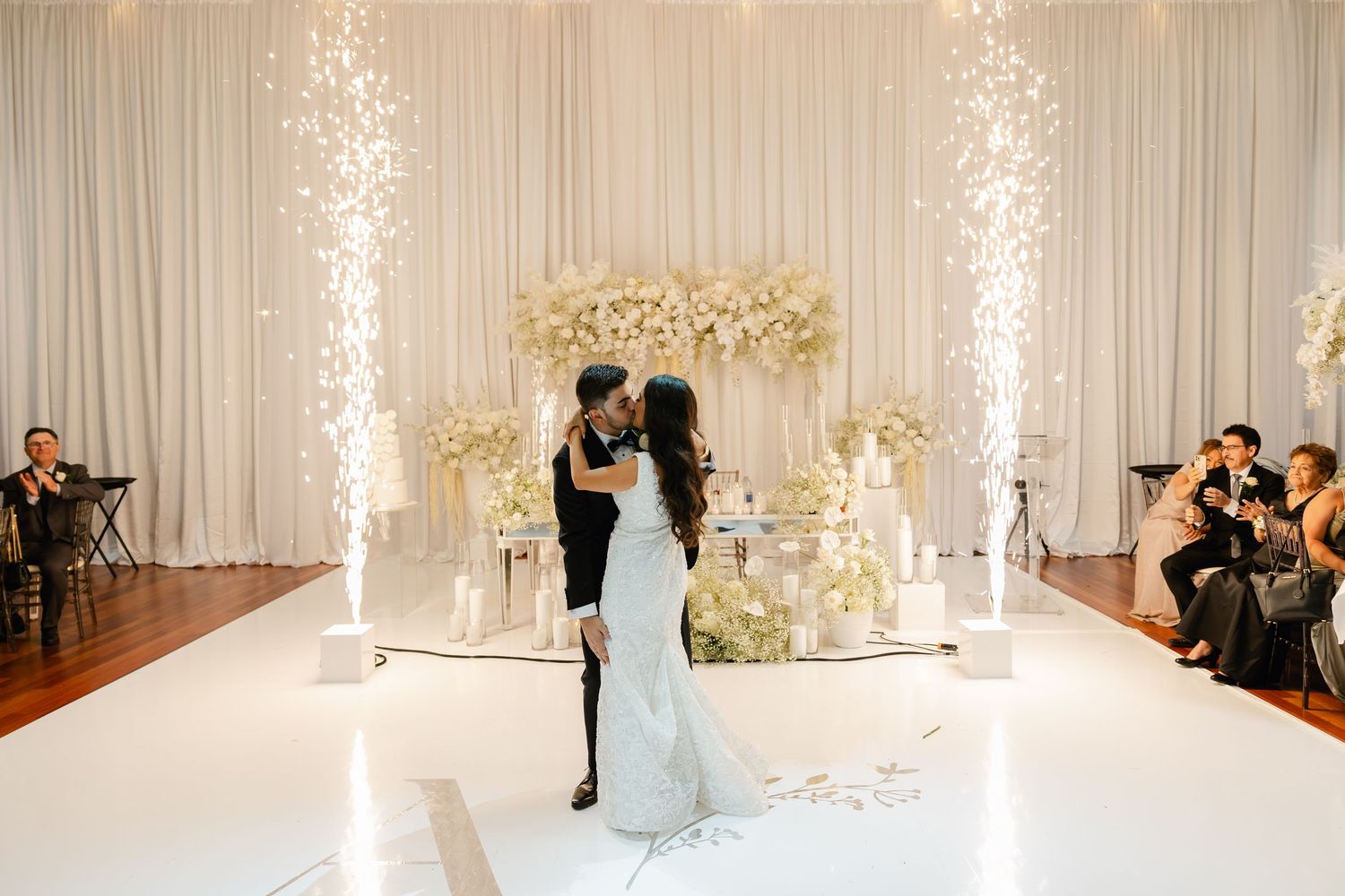 Wedding couple dancing on white platform with sparkler fountains and floral backdrop.