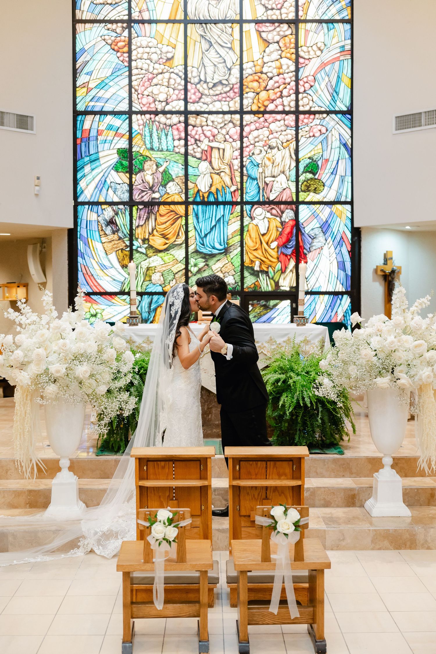 Wedding ceremony with beautiful stained glass backdrop and white flower decorations in a church setting.