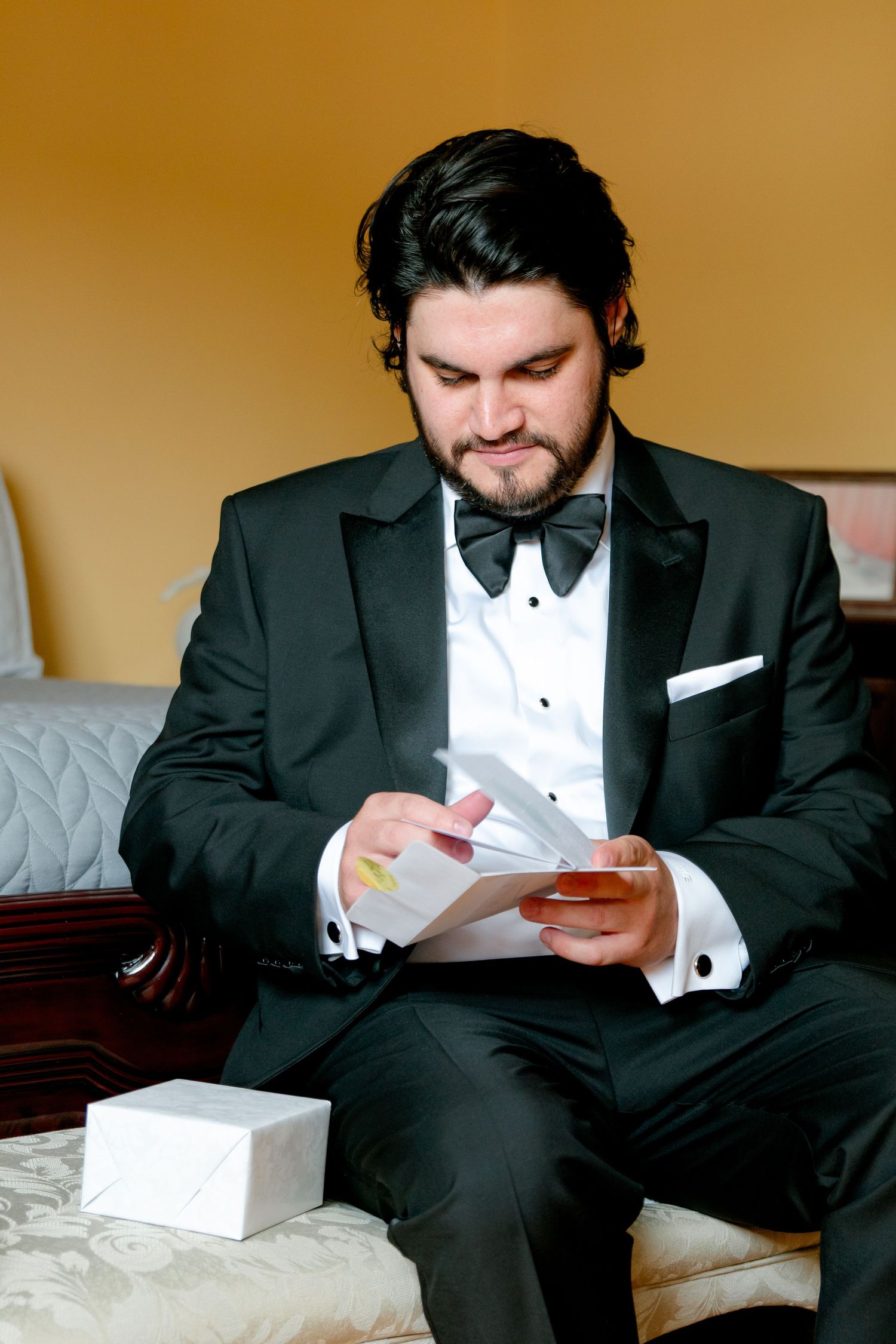 Man in black tuxedo sitting on bed reading a note or card with thoughtful expression.
