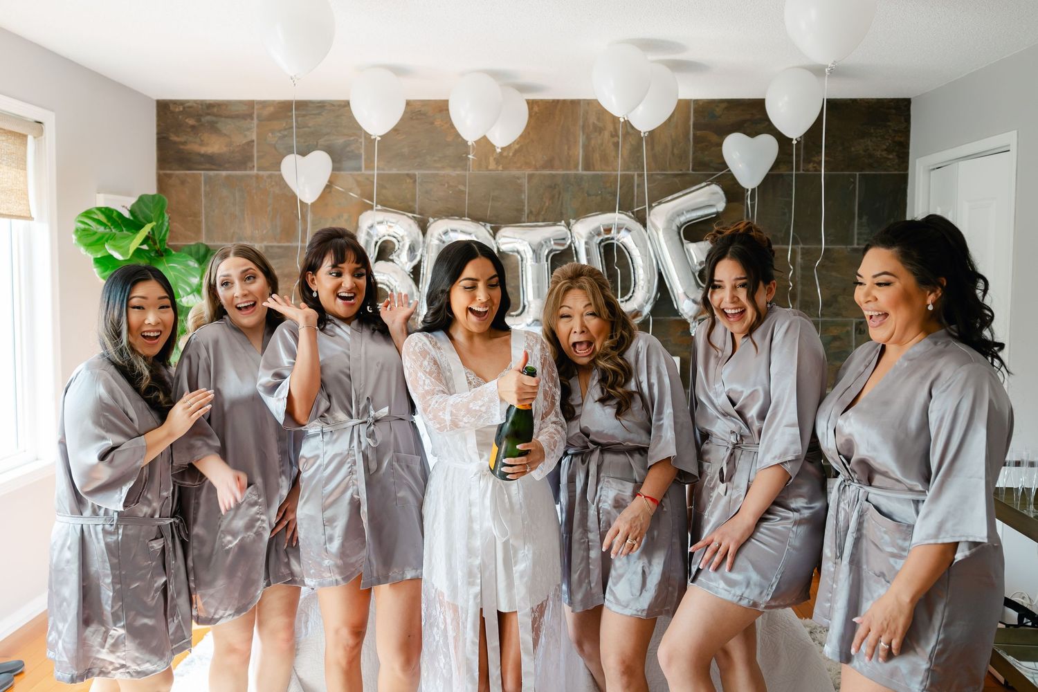 Wedding party in matching silk robes poses together with champagne under white heart balloons.