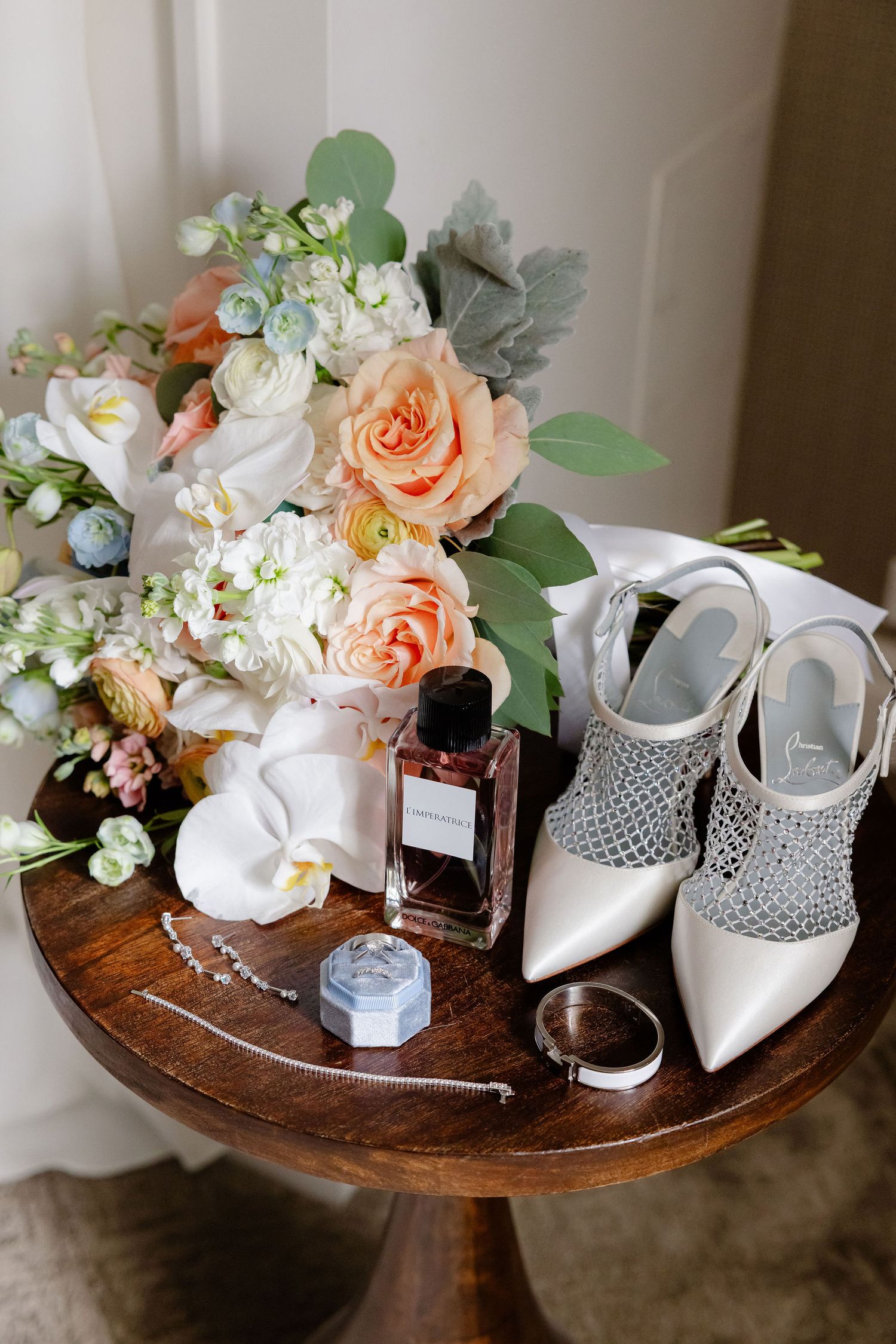 Wedding accessories including elegant white shoes, jewelry, and peach floral arrangements displayed on a wooden table.