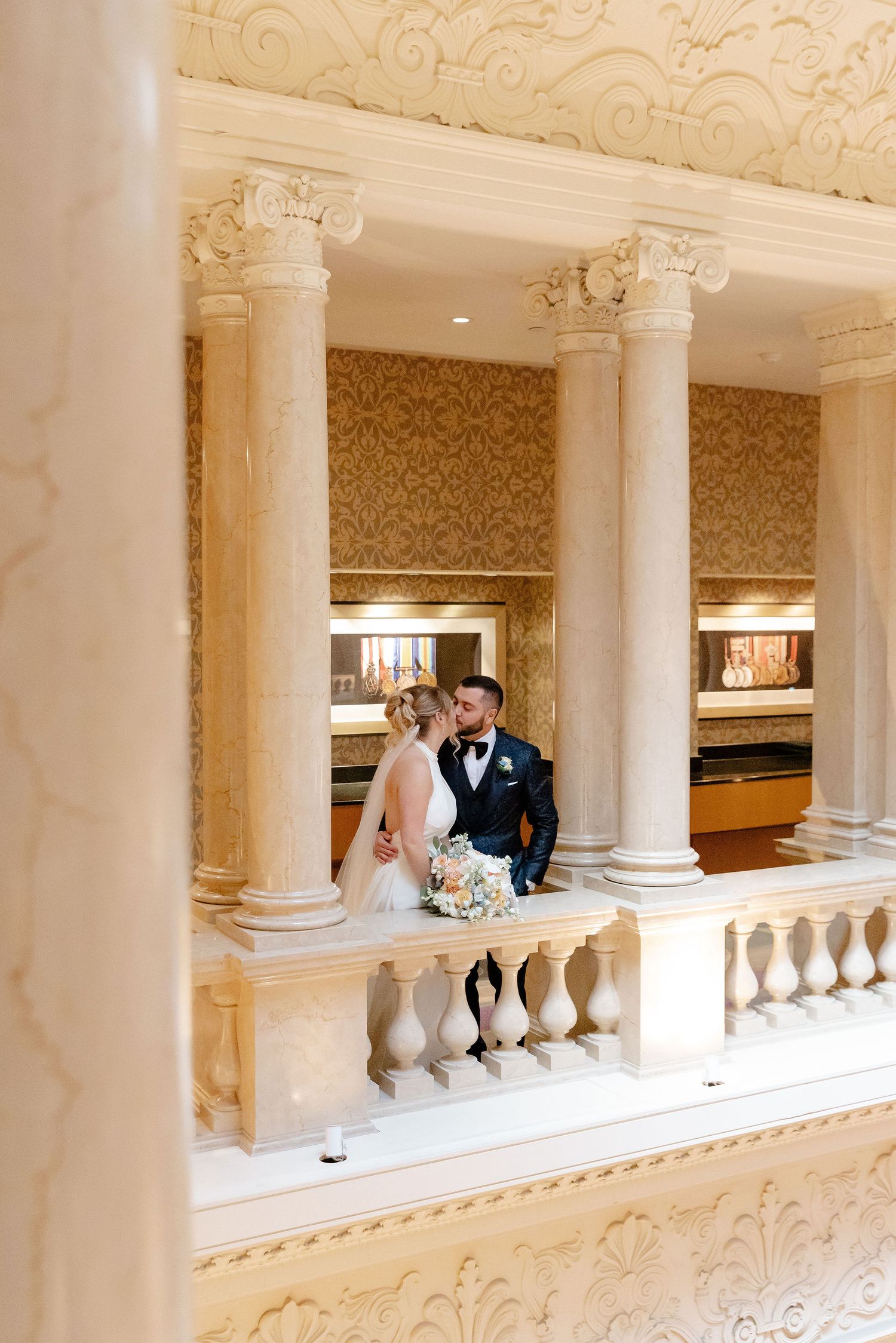 A wedding couple poses between classic white columns along an elegant interior balcony.