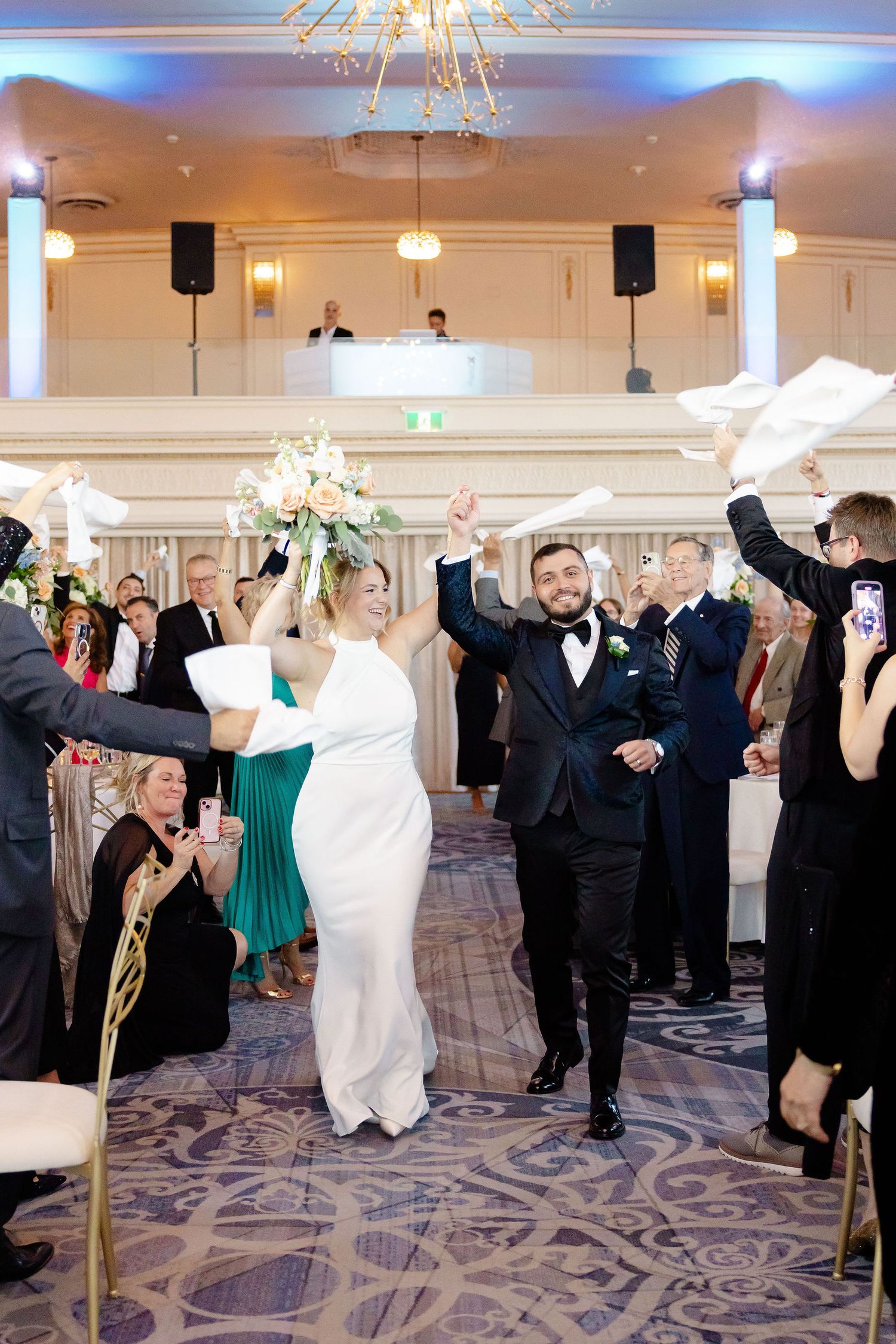 Wedding guests dancing and celebrating in an elegant ballroom with ornate carpet and chandeliers.