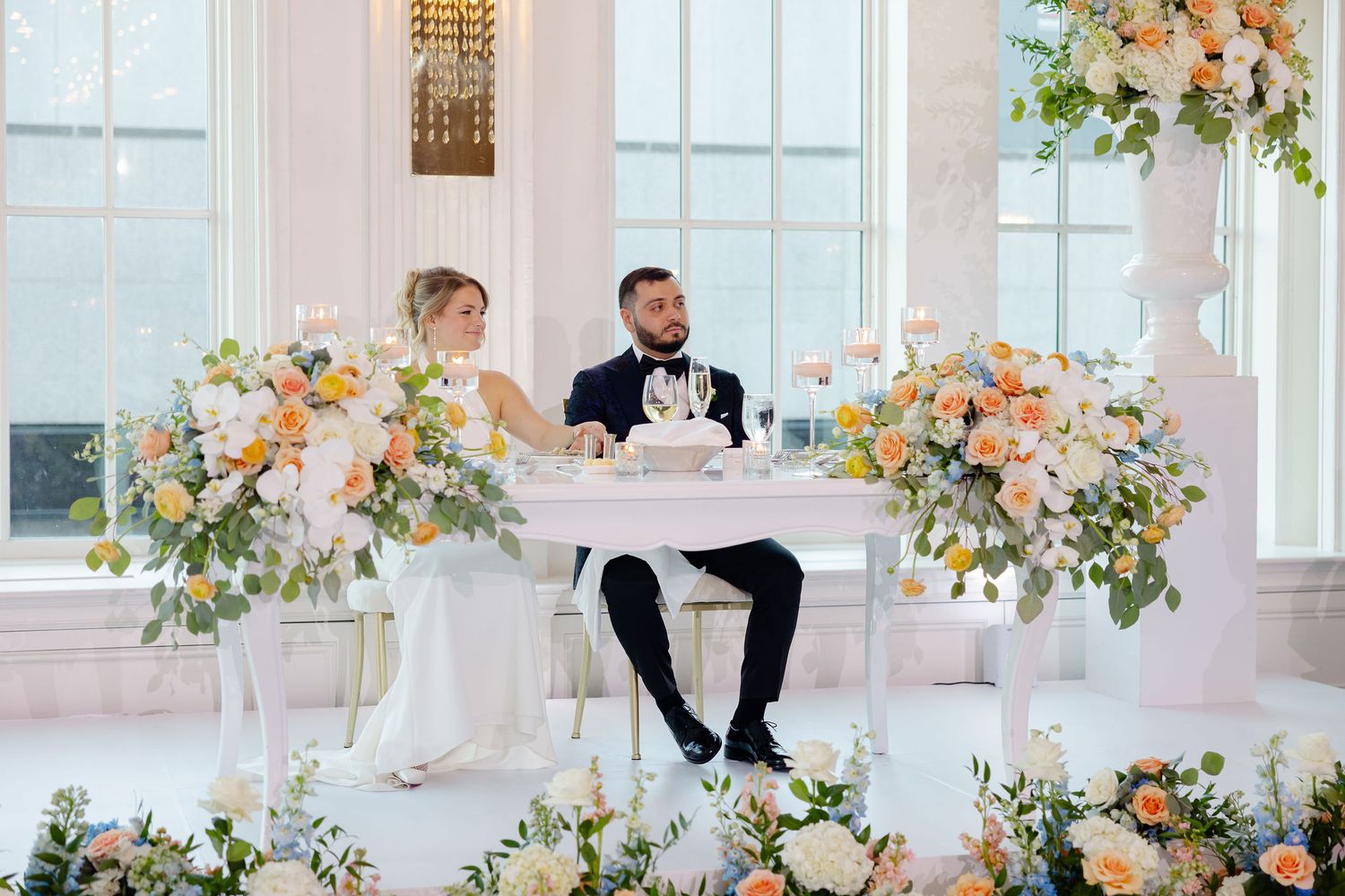 Wedding couple at reception venue with elegant floral arrangements and large windows.