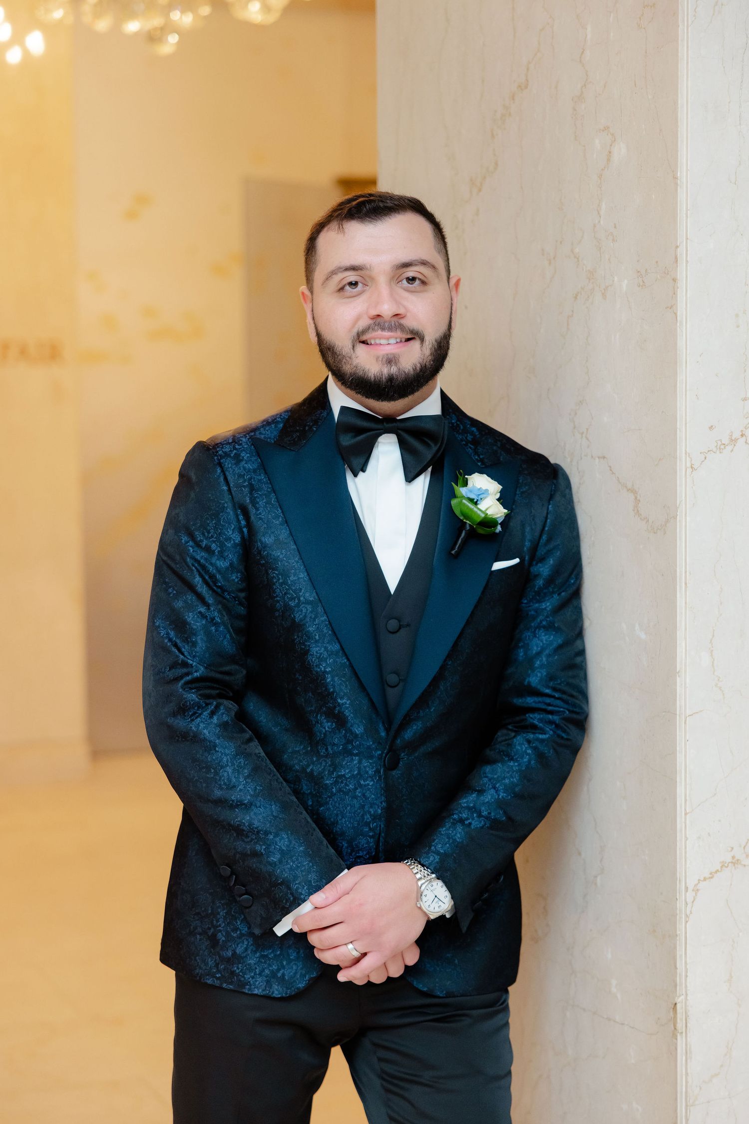 Groom in navy tuxedo poses against marble walls in an elegant venue with warm lighting.
