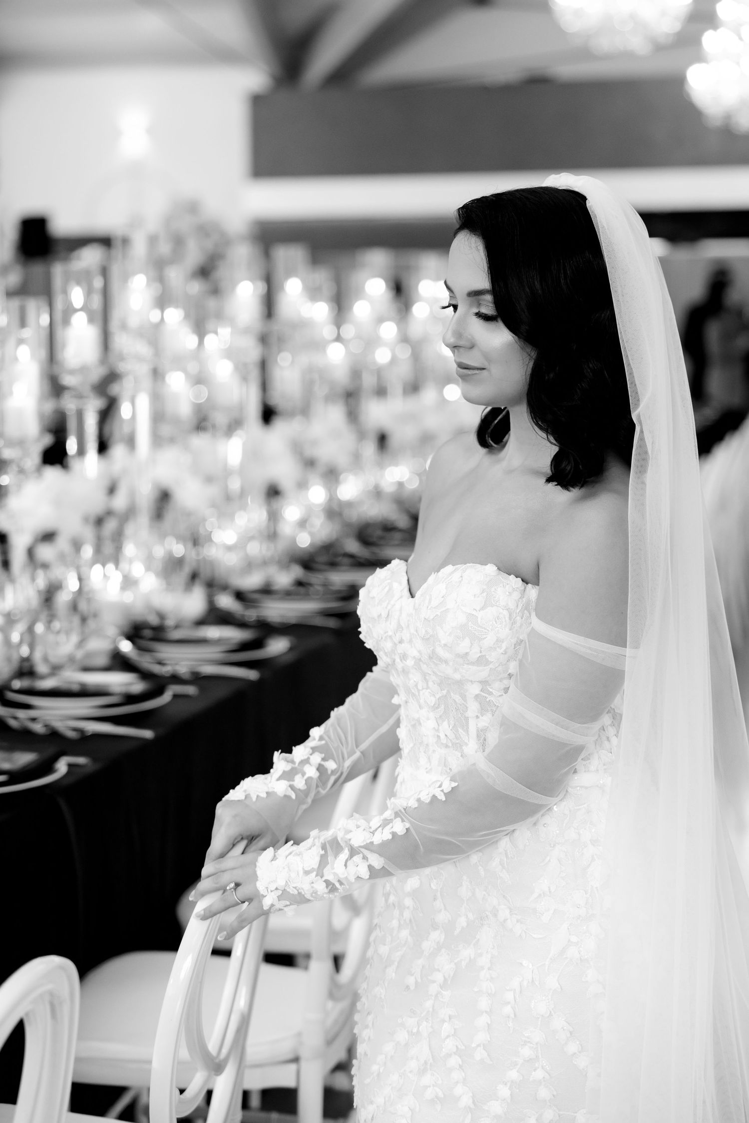 A black and white photo featuring a bride in a lace gown seated at an elegantly set wedding table.