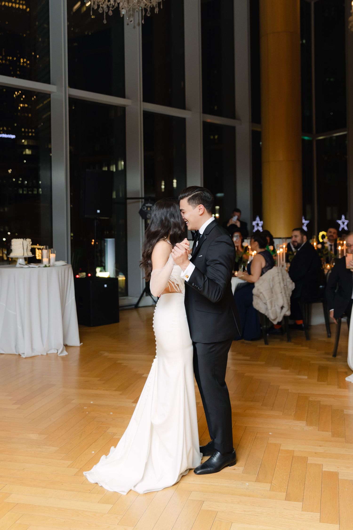 Newlyweds dancing in a modern reception venue with chandelier and city views.