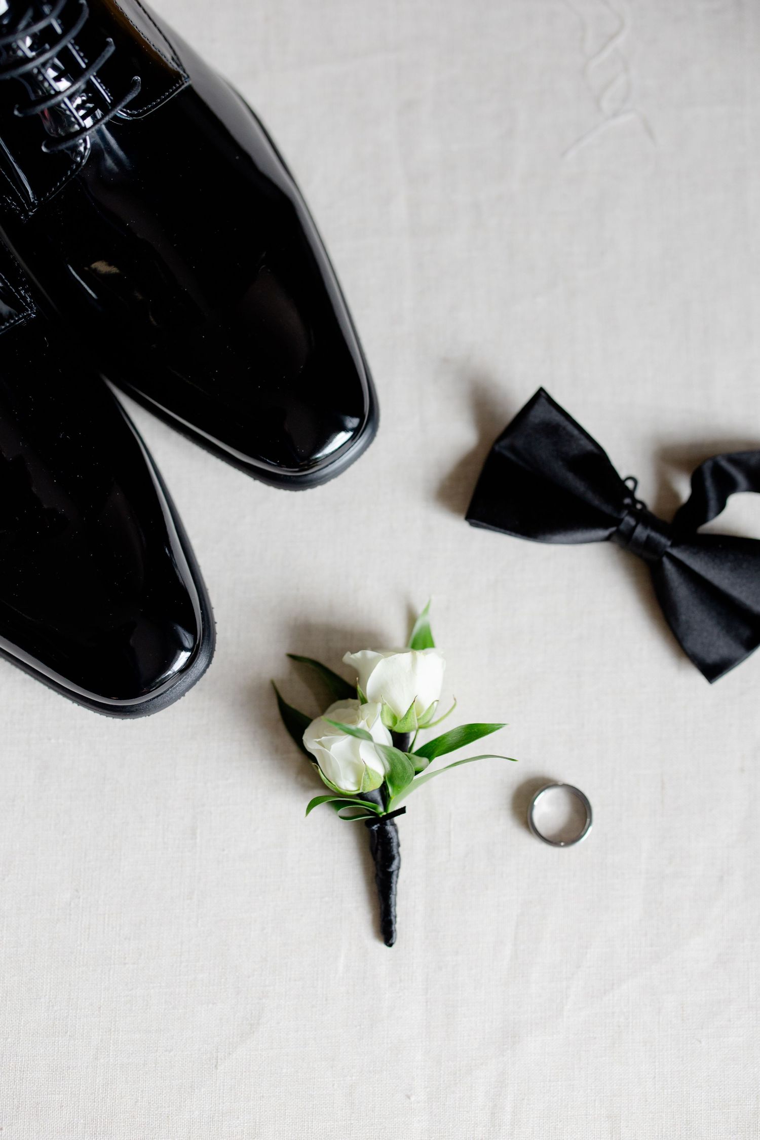 Wedding accessories including black dress shoes, bow tie, boutonniere and ring on white background.