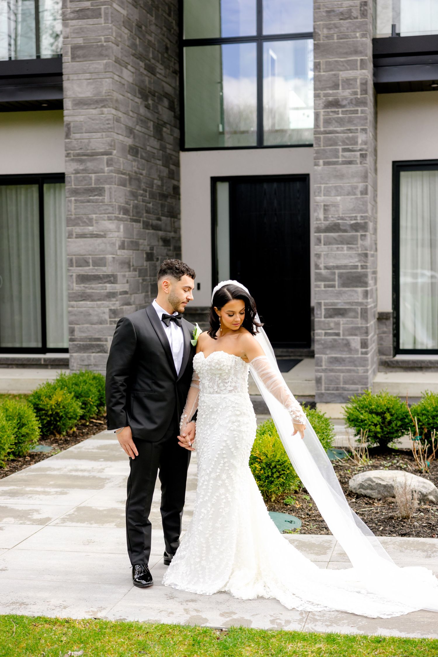 A wedding couple strolls hand in hand along a pathway with a flowing white veil trailing behind.