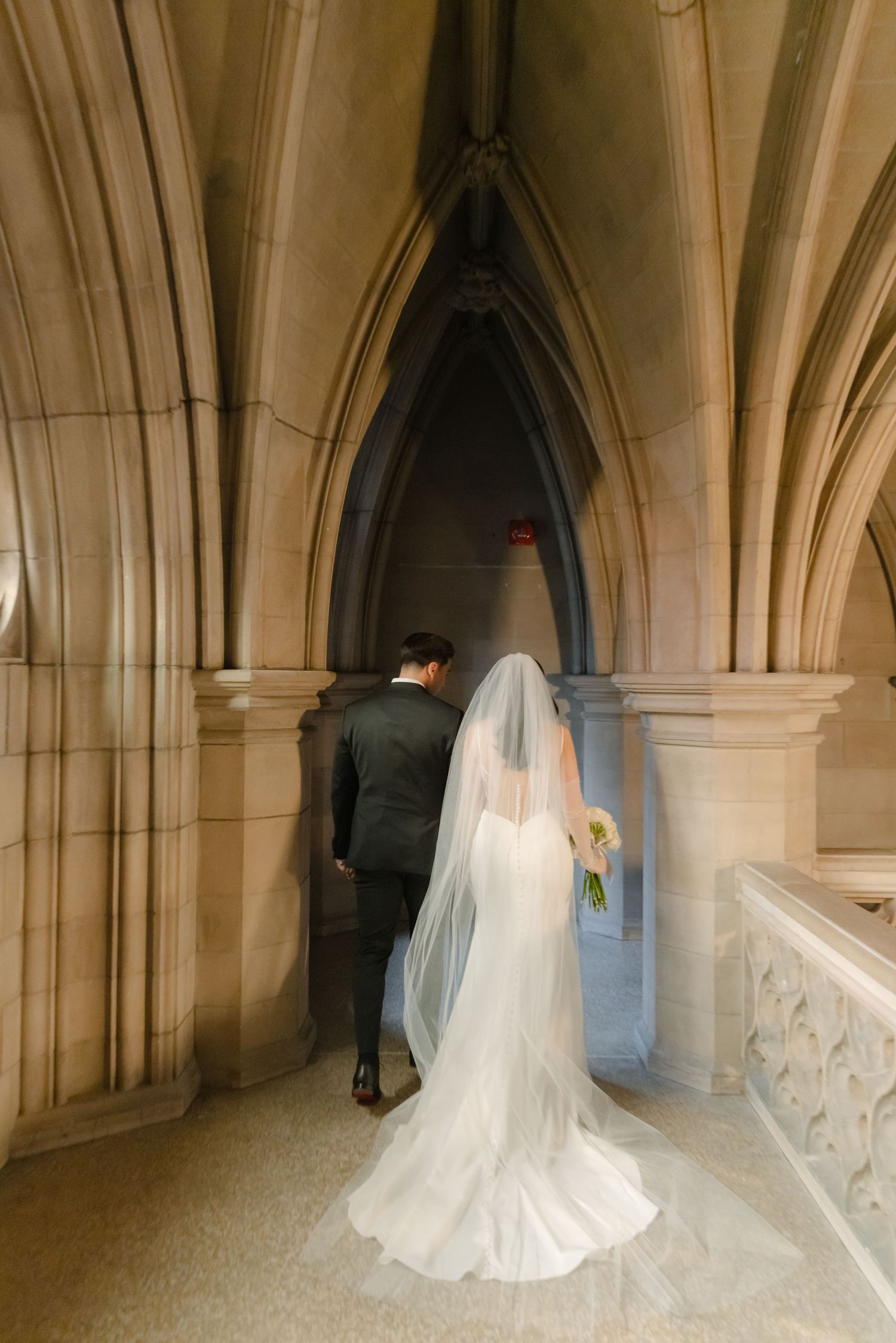 A wedding couple walks through a gothic stone corridor with dramatic vaulted ceilings.
