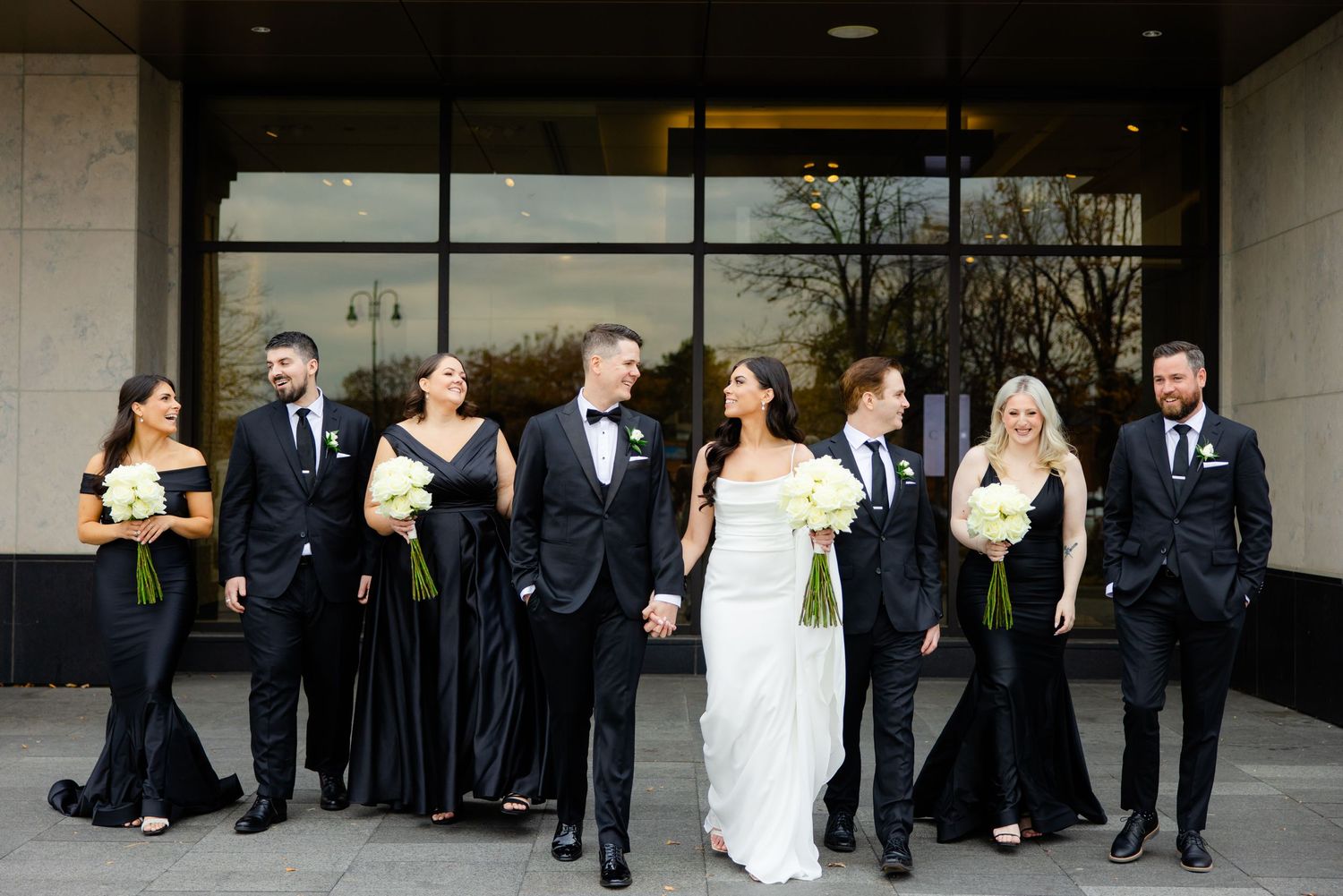 Wedding party in formal attire walks together outside modern building entrance.