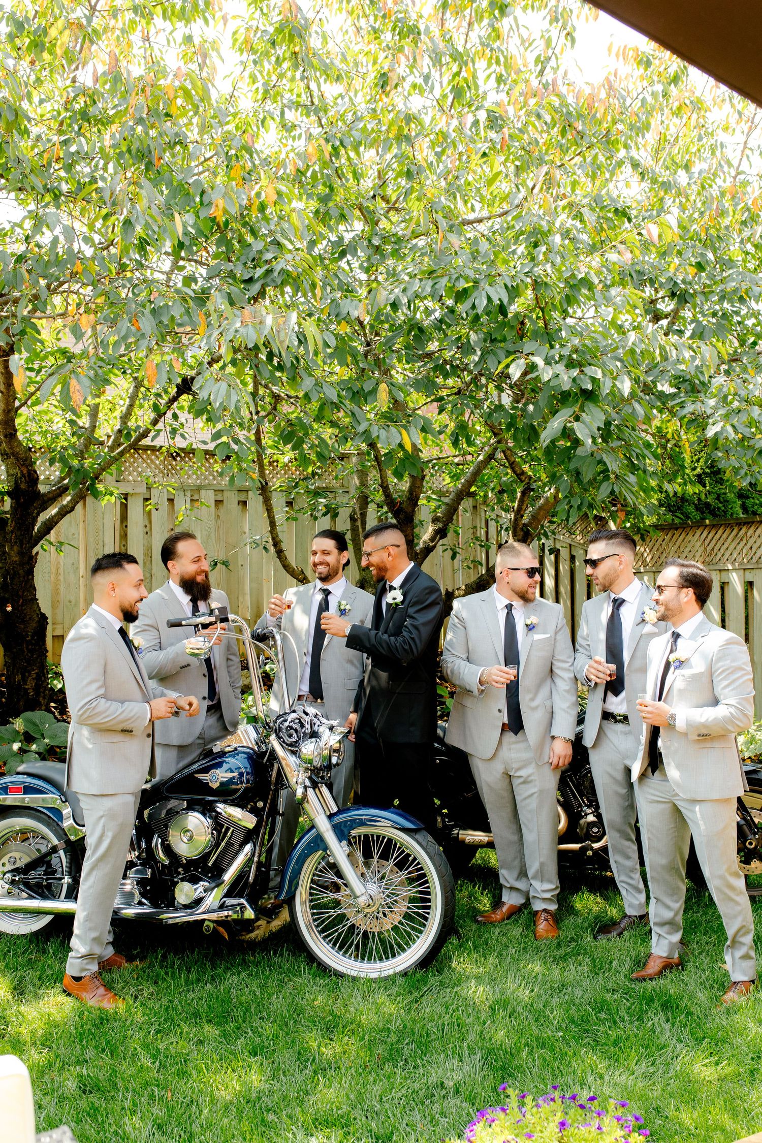 Groomsmen in light suits sharing drinks around classic motorcycles in a garden setting.