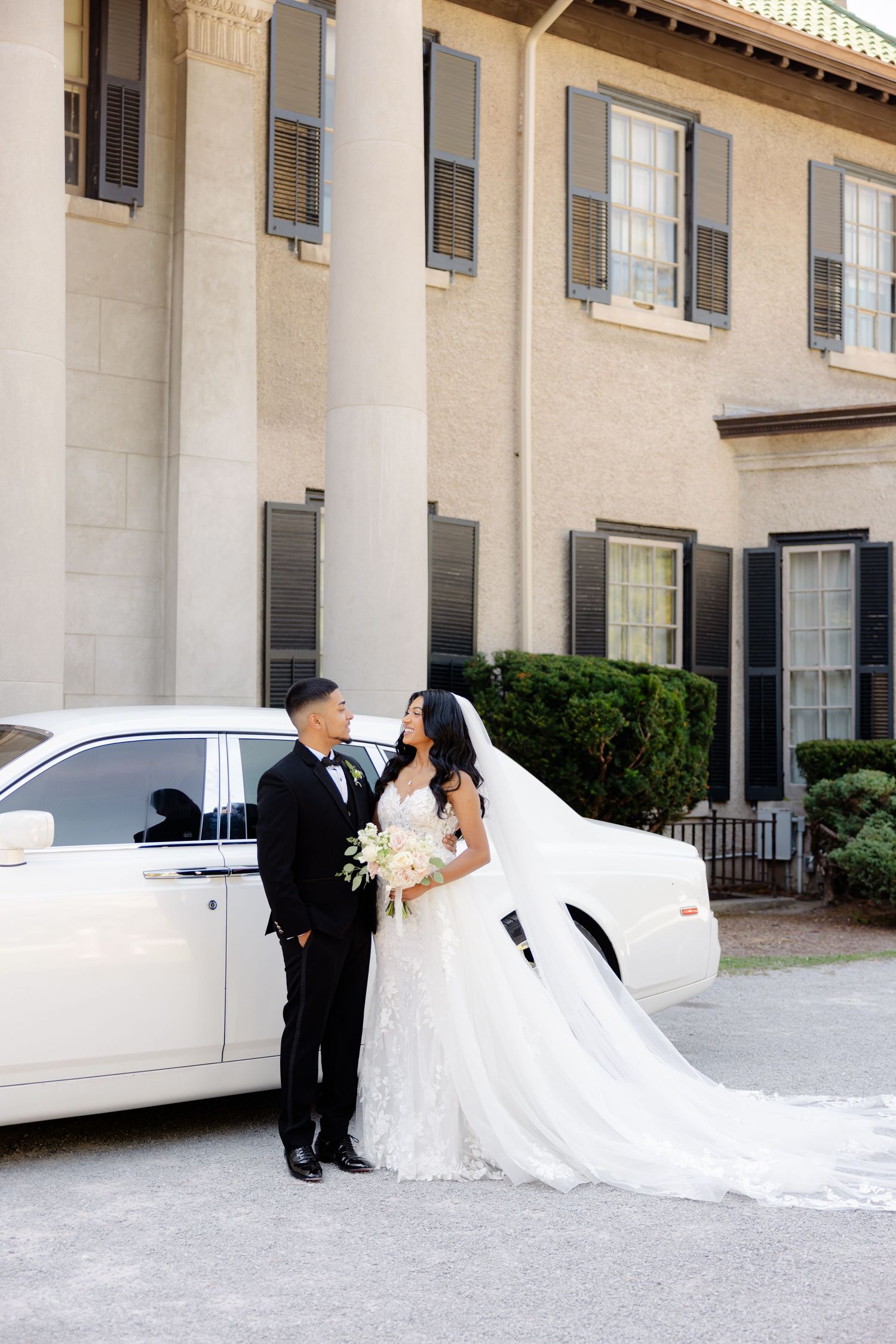 Elegant wedding portrait with a couple standing by a white luxury car outside a stately building.