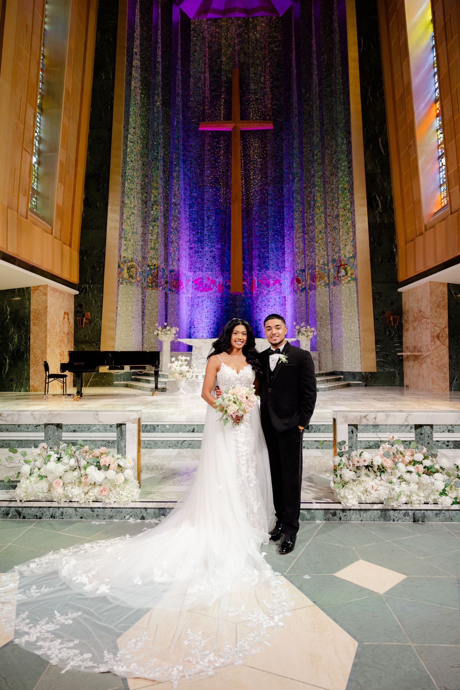 A wedding couple poses in front of a dramatic purple-lit church altar with a large cross and floral arrangements.