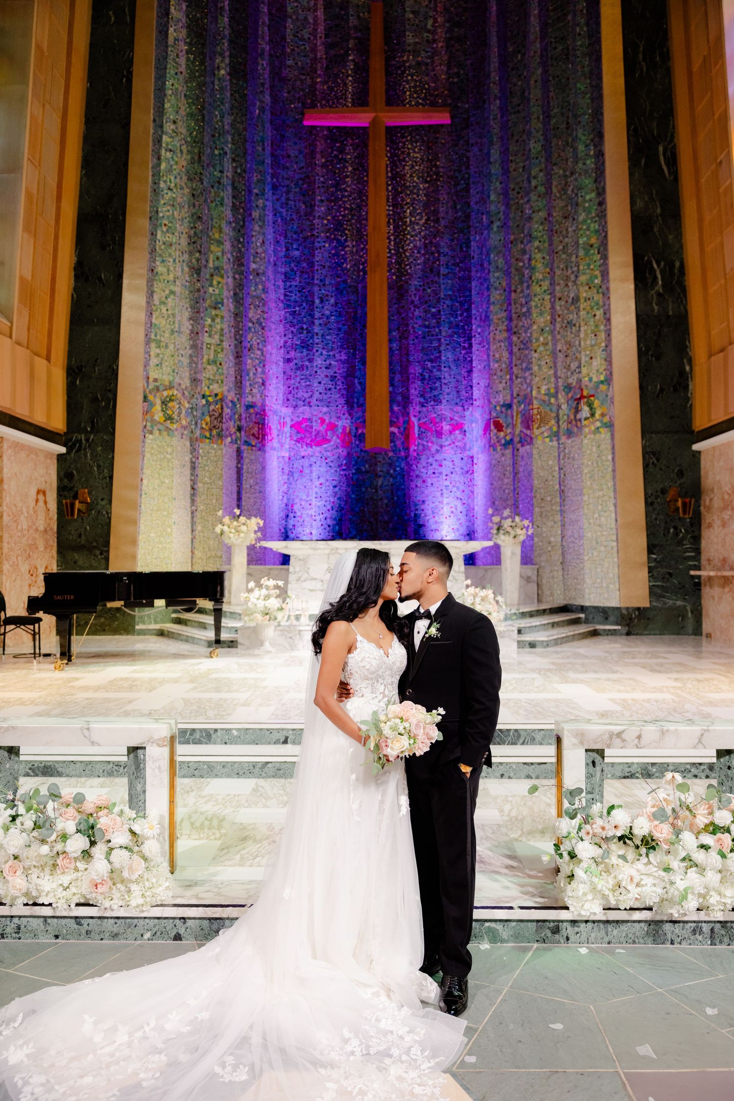 Romantic wedding photo with couple standing before illuminated cross and purple church backdrop.
